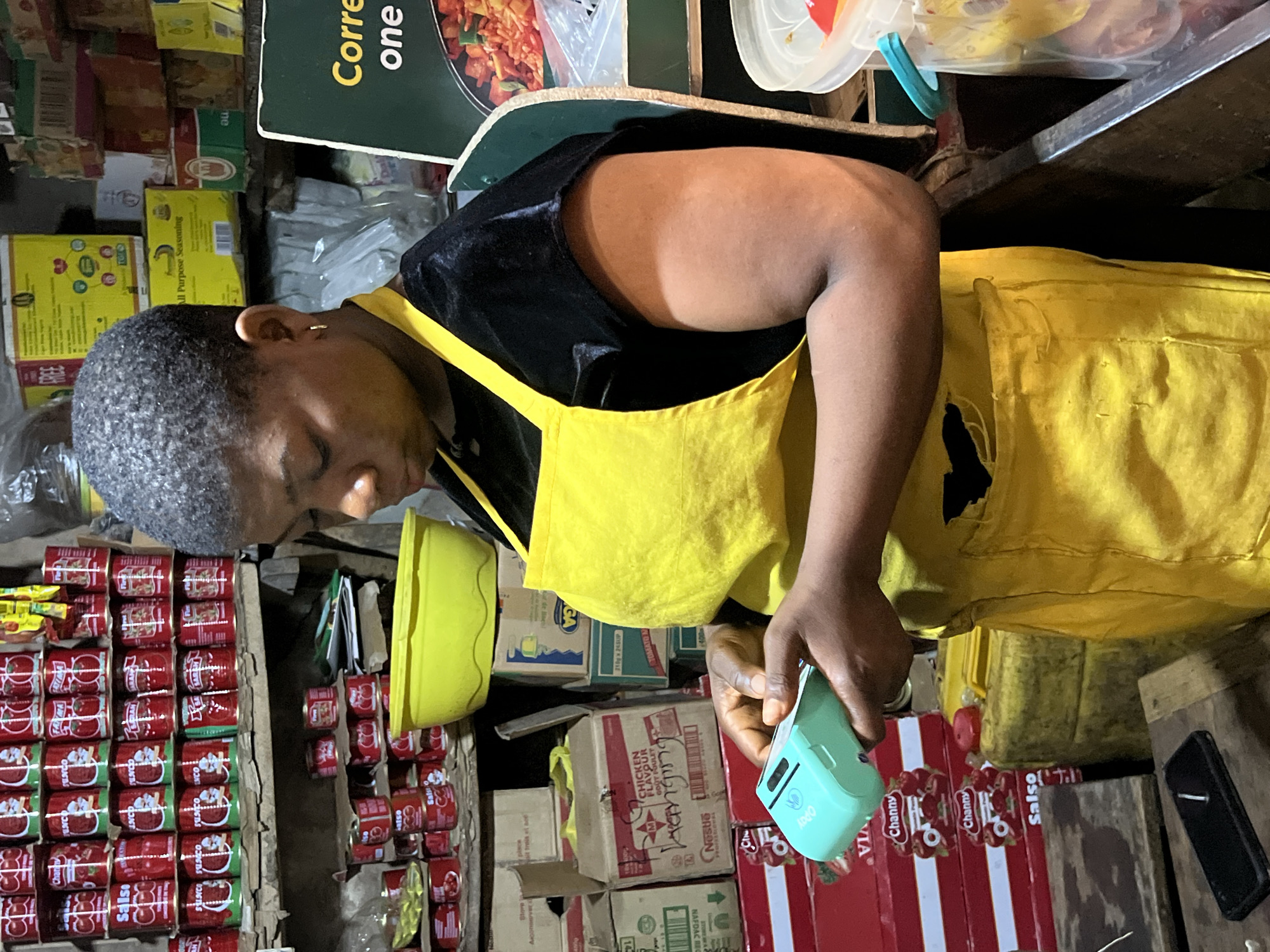 The image shows a woman in Nkpor, Nigeria, standing in what appears to be a small retail space. She is wearing a yellow apron over a black top. In her hand, she is holding a small, light-blue device, possibly a barcode scanner or similar handheld electronic device.
The background is filled with a variety of canned goods, specifically "Salsko" tomato paste cans, along with boxes of "Channy" tomato paste and other products such as "All Purpose Seasoning." There are also boxes of "Chicken Flavour" and "Nestle" products, further indicating a retail environment. The overall space seems cramped, with goods stacked tightly together. A green sign with white writing and a food image is partially visible at the top of the image.