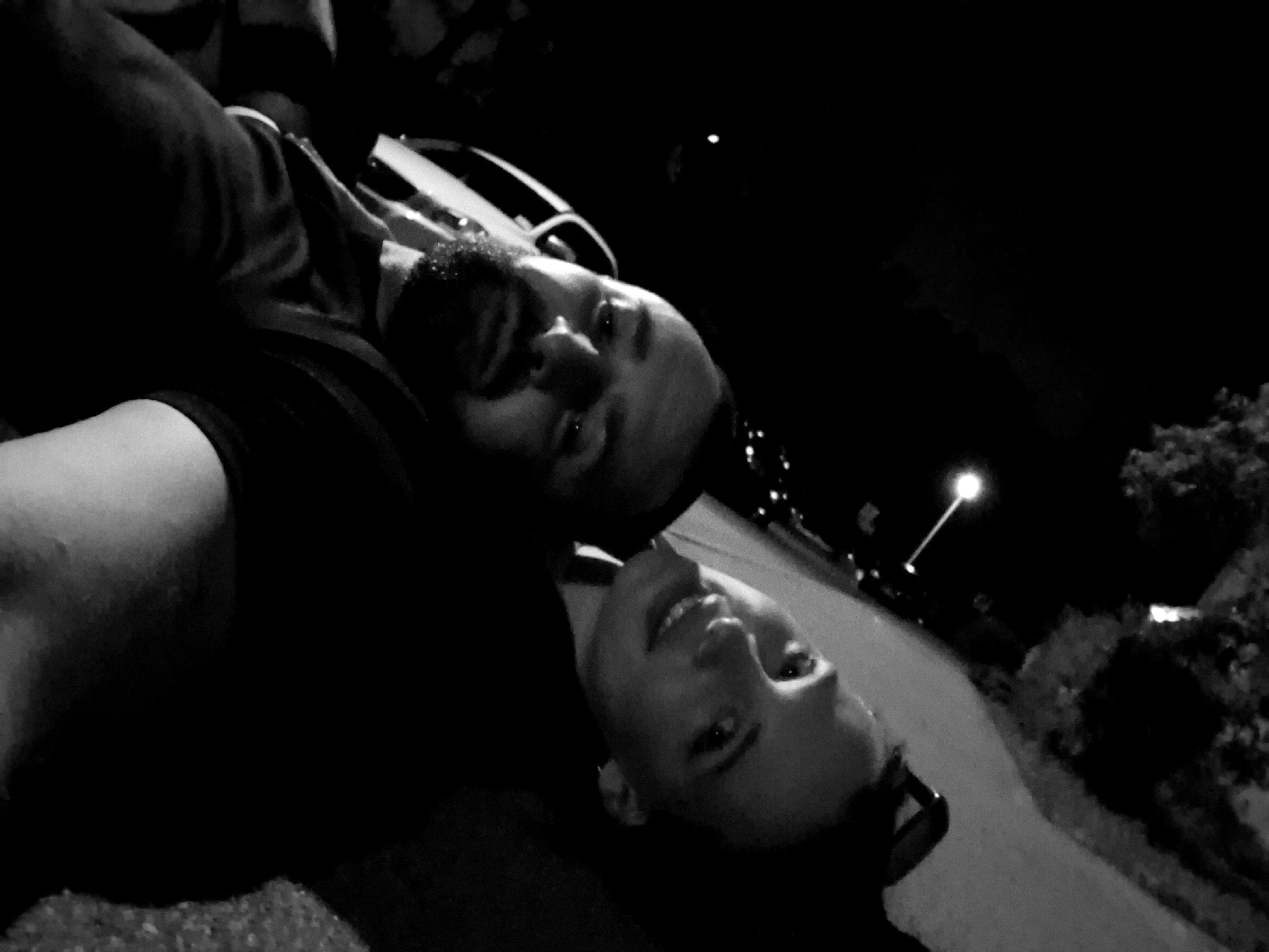 In a black and white, low-light, outdoor image, two individuals are lying down, facing towards the viewer, with their heads close together. The person on the left, who appears to be male, has a beard and is looking directly at the camera with a slight smile. The person on the right, who appears to be female, is looking up and to the left with an open-mouthed smile. The individuals are lying on a surface that appears to be gravel or rough pavement. In the background, a street or path is visible, with a light pole and some trees or foliage. The scene suggests a casual or intimate moment captured at night.