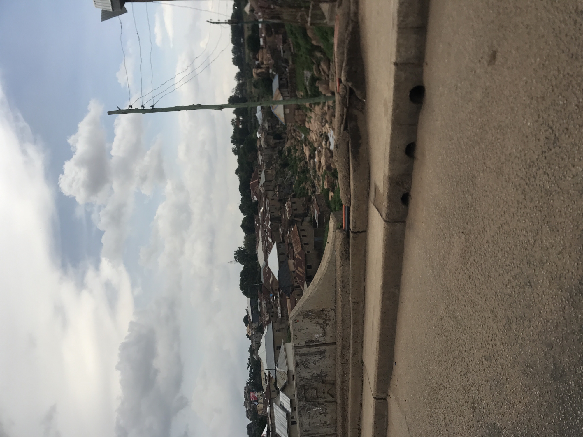 This image captures a scene in Jos, Nigeria, depicting a densely populated urban landscape characterized by varied architectural styles and a rocky terrain. In the foreground, a paved road with a concrete curb and drainage openings is visible, suggesting infrastructure within the city. The middle ground features a concrete barrier, possibly part of a bridge or overpass, overlooking a collection of buildings. These structures are predominantly single-story, with many exhibiting corrugated metal roofs, some appearing rusted. The buildings are tightly packed, clustered on a hillside and interspersed with rocky outcrops. Vegetation, including trees, is present on the slopes and around the buildings. In the background, a cloudy sky with patches of blue dominates the scene. A wooden utility pole with electrical wires stretches vertically through the frame, indicating the presence of power lines. The overall impression is of a vibrant, working-class urban environment situated in a naturally rugged setting.