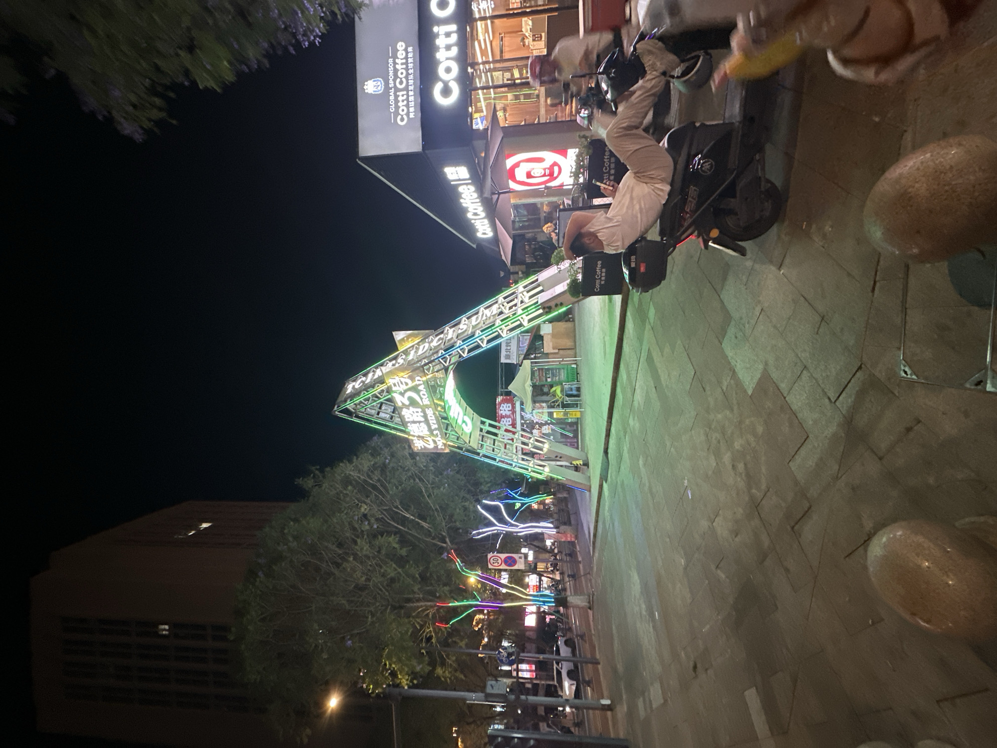 The image depicts a street scene at night in what appears to be the Chongqing High-tech Zone in China. The scene includes street lighting, a brightly lit Cotti Coffee shop with signage in English and Chinese, and a large decorative archway with the Chinese characters "艺德路3号 NO.3 YIDE ROAD". There are trees decorated with colorful lights along the street. A person in light-colored clothing is sitting on a parked scooter or motorcycle near the coffee shop. Additionally, there's a sign indicating Cotti Coffee as a global sponsor of the Argentinian national football team. The street is paved with what looks like a tile pattern. A building can be seen in the background.