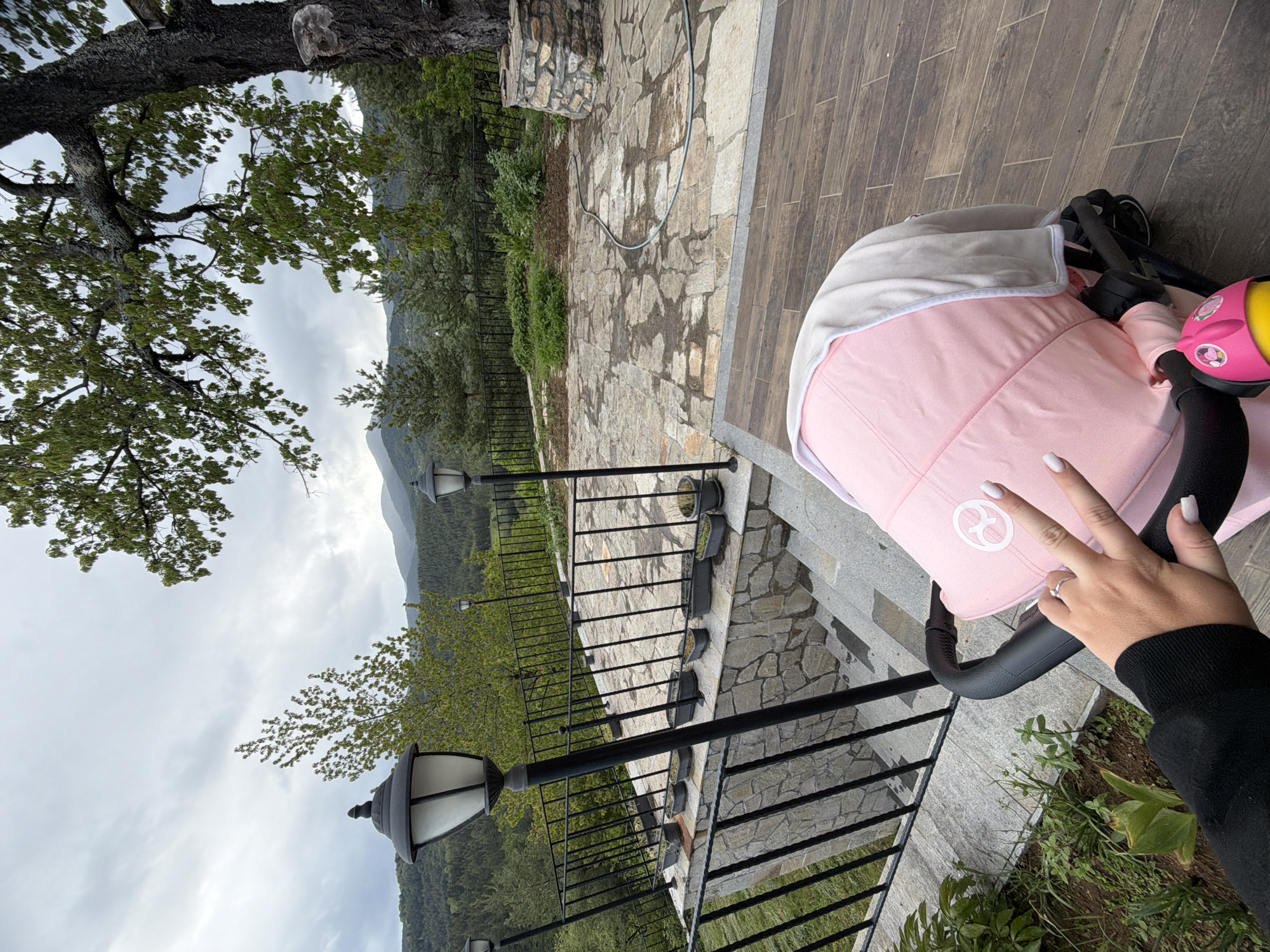 A high-angle shot shows a pink baby stroller with a person's hand displaying a peace sign on a patio in Кирково, Bulgaria. The patio is composed of stone tiles and wood planks, bordered by a black iron railing. Behind the railing, a hillside is covered in green trees and vegetation, with distant mountains visible under a cloudy sky. A large tree with thick branches and green leaves is visible in the top left corner of the frame. A black sweater sleeve is visible on the right side of the hand holding the peace sign.
