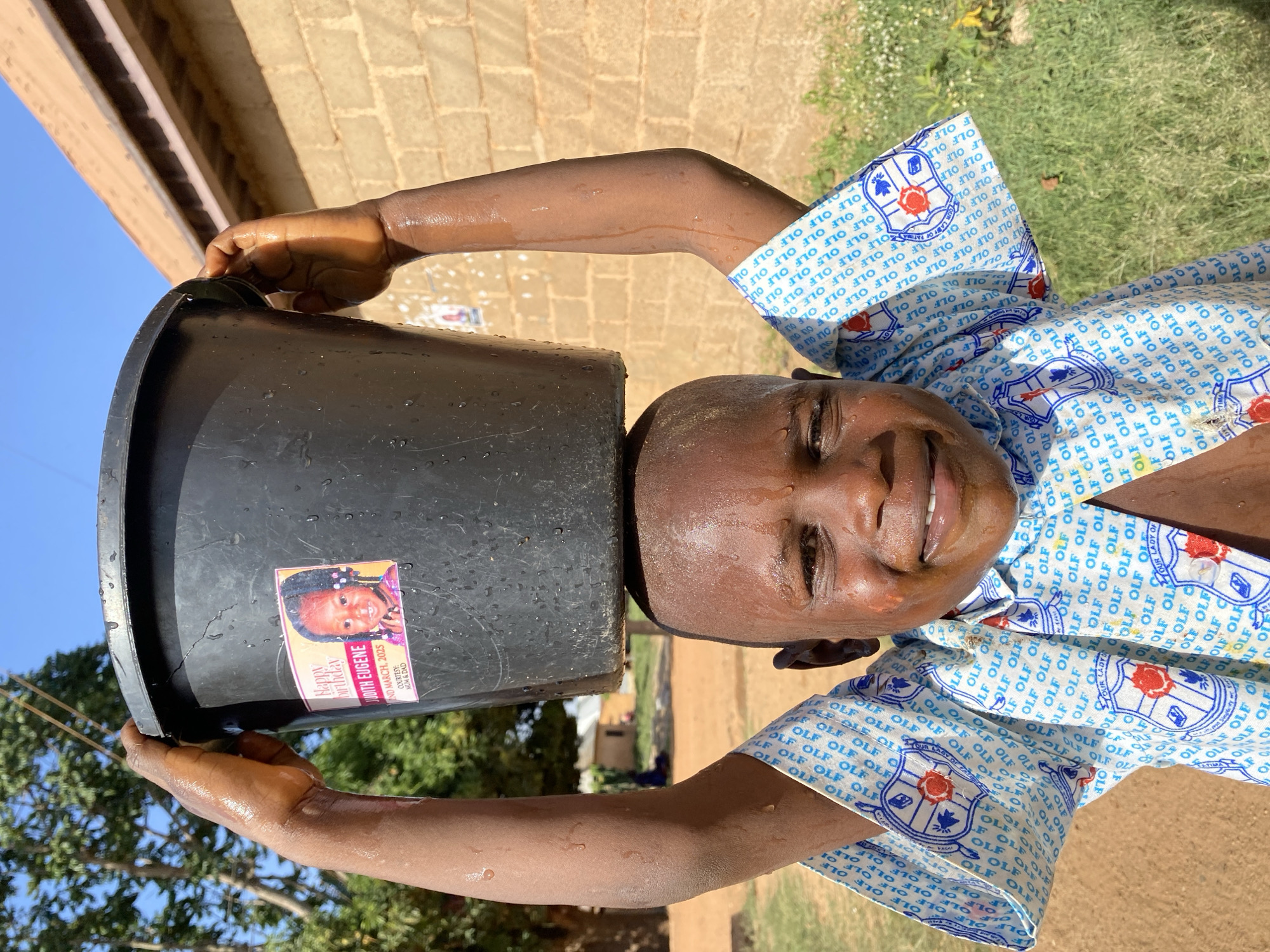 A young, dark-skinned child smiles with a black bucket on their head. They are outside, in the Kuru Dungus area of Nigeria, near a brick wall and vegetation. The child's arms are raised, holding the bucket in place. The bucket has a sticker on it with a picture of a young girl with the words "Happy Birthday Judith Eugene 2nd March 2025". The child is wearing a blue and white patterned shirt with “OLF” printed on it repeatedly. The child's face and arms are wet.
