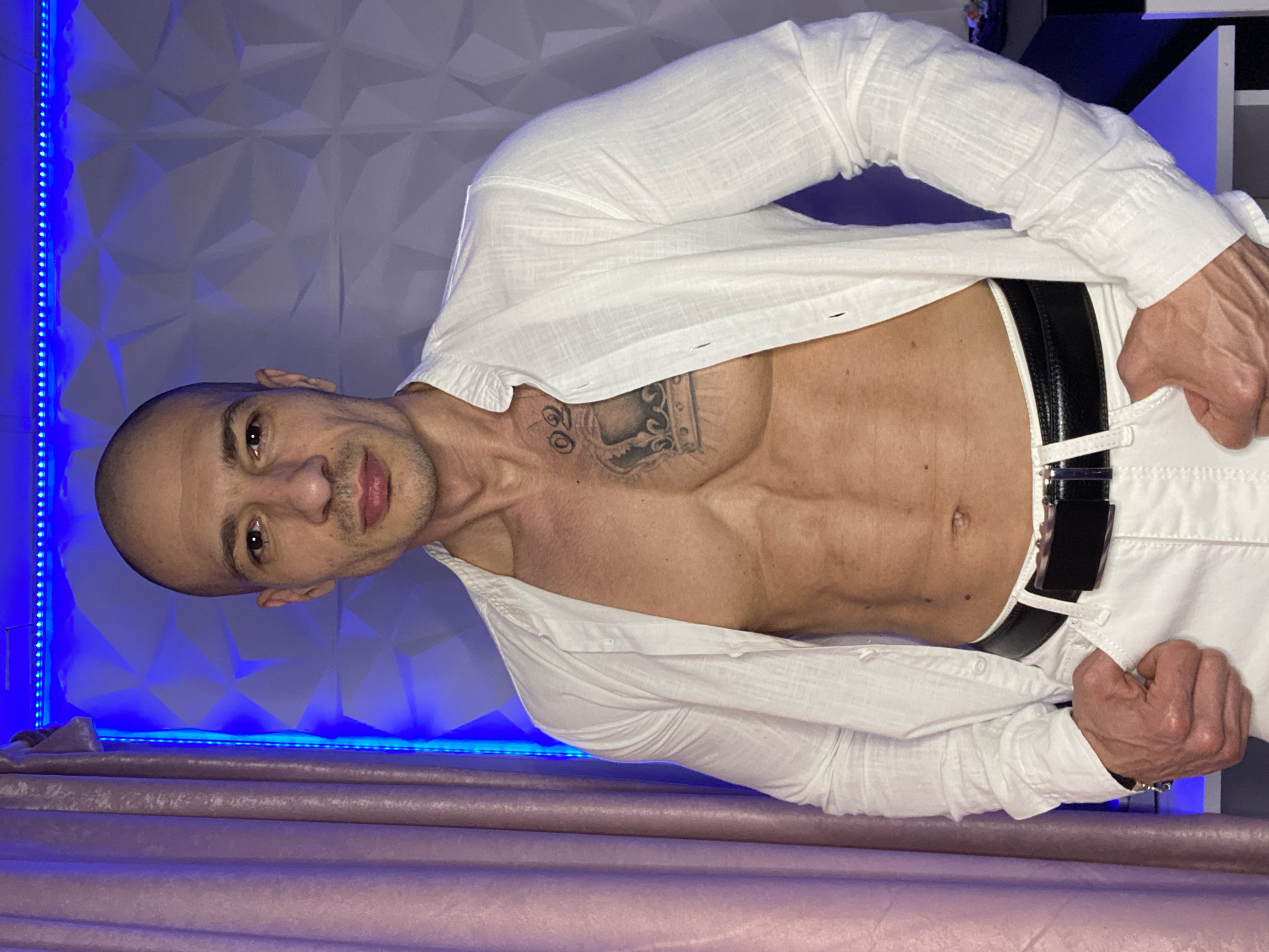 An adult male stands facing forward, making direct eye contact with the viewer. He has a bald or closely-shaven head, a short beard or stubble, and a muscular build with visible abdominal definition. He is wearing an unbuttoned white long-sleeved shirt, exposing his torso, white trousers, and a black belt with a silver-colored buckle. A tattoo of a crown with the numbers "02" directly above it is present on his upper left pectoral. Both hands are partially inserted into the pockets of his trousers. The background consists of an indoor setting with a light pink/purple curtain on the left and a white geometric 3D textured wall on the right. A strip of blue LED lighting runs horizontally along the top edge of the textured wall. The specific location within the city of Pleven, Bulgaria, cannot be determined from the visual information provided.