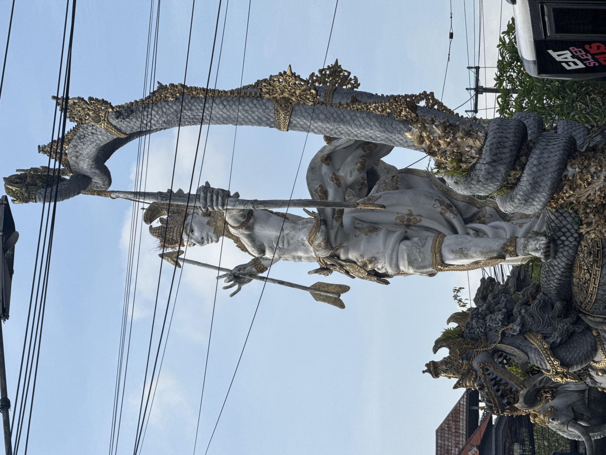A large, ornate stone statue stands prominently against a bright, partly cloudy sky. The central figure, likely a deity or hero, is depicted in a dynamic pose, aiming a bow with an arrow nocked. This archer statue is carved from light grey stone and adorned with intricate gold accents on its elaborate headwear, armbands, and clothing.
Behind and partially encircling the archer, a magnificent, scaled serpent or dragon-like creature forms a grand arch, its body detailed with grey scales and rich gold ornamentation. The head of this mythical creature, with fierce teeth and ornate features, is visible at the lower right of the statue, forming part of its detailed base. The overall style of the sculpture, combining a heroic figure with a naga-like creature and intricate gold detailing, is characteristic of traditional Balinese art, consistent with its location in the Ubud District, Indonesia.
Numerous black utility cables crisscross the upper and middle sections of the frame, passing in front of the statue and against the light blue sky. To the far right, green foliage from trees or bushes is visible, and below it, a reddish-brown building roof can be seen. In the top right corner, a partial view of a dark-colored vehicle or sign displays white text that reads "EAT", "SLEEP", and "RIDE". The bright sky indicates it is daytime with fair weather.