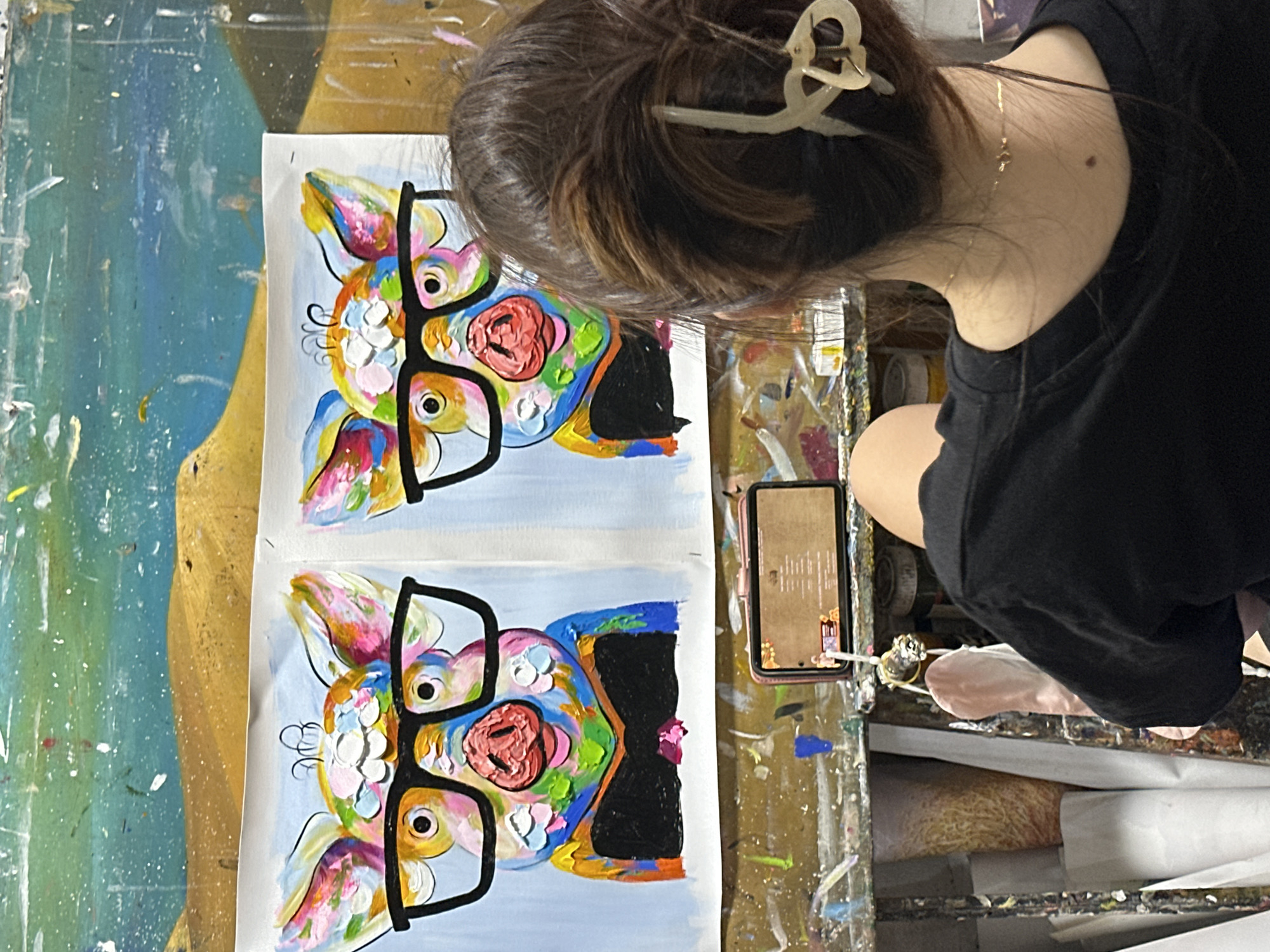 From a high-angle perspective, the scene presents a person working on a colorful painting on a possibly cluttered workbench. The individual, wearing a black t-shirt, has their dark brown hair pulled up with a light-colored hair clip. A gold necklace is visible on the back of their neck. Two identical paintings, depicting a colorful animal portrait with large black glasses, sit side-by-side on white paper. A smartphone rests on the workbench beside the paintings, possibly displaying reference material or instructions. The bench is stained with paint and debris. I am unable to provide the exact location in the city of Thủ Đức, Vietnam.