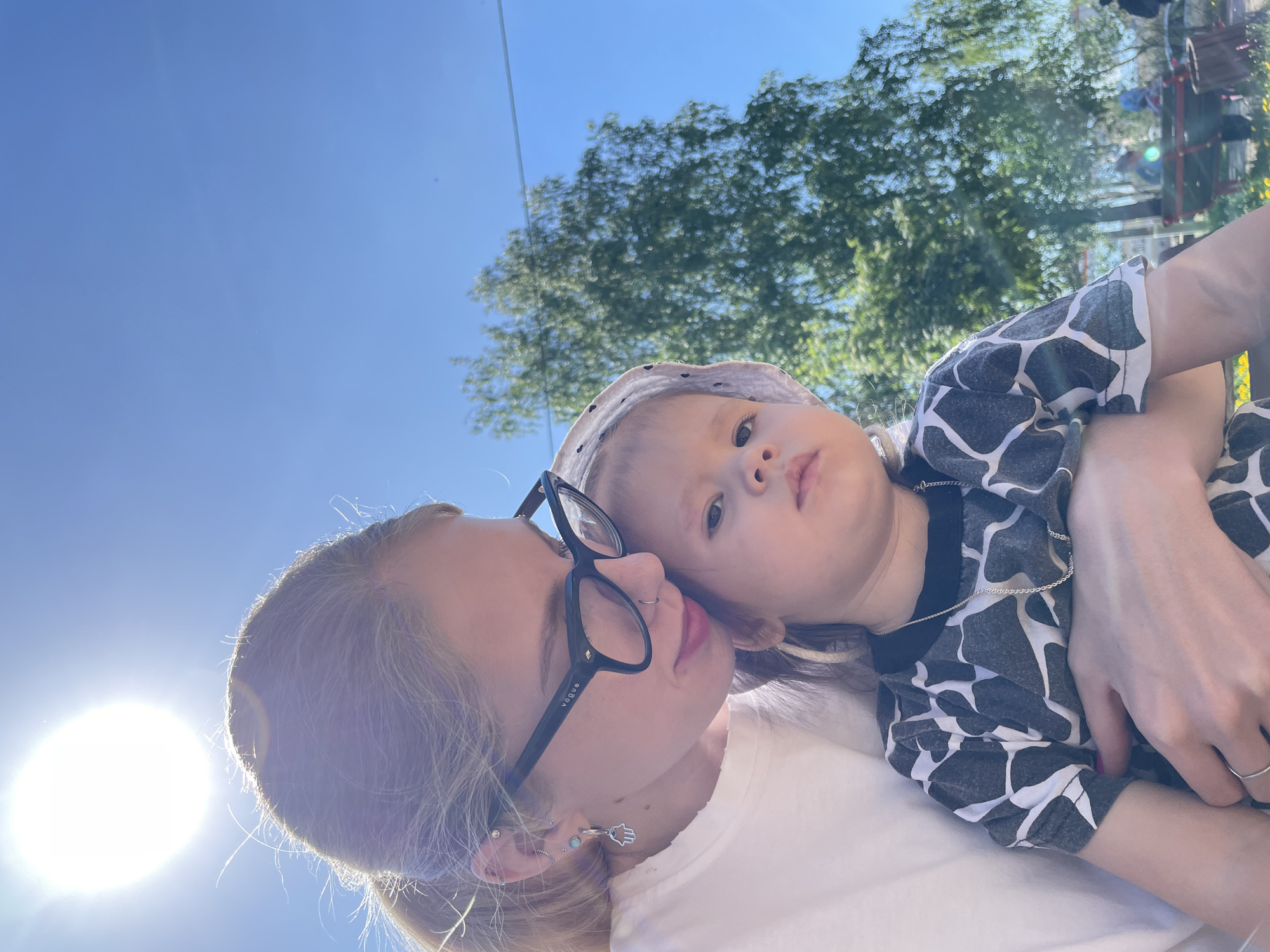 A woman wearing black-rimmed glasses and a white t-shirt is holding a young child wearing a spotted grey and white shirt and a dotted hat. The background includes a clear blue sky, a leafy tree, and a blurred playground structure. The woman appears to be taking a selfie. Based on the playground equipment and the clothing of the individuals, it's most likely a park or recreational area in Izhevsk, Russia.