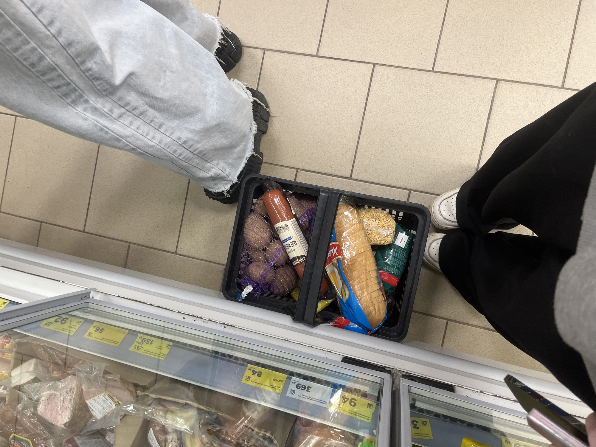 The image shows a top-down view of a shopping basket filled with groceries, standing in front of a refrigerated display case in what appears to be a grocery store. Two people are visible; one wearing light-colored jeans and black shoes, the other wearing black pants and white shoes. The basket contains potatoes, a sausage (possibly labeled with Cyrillic text), bread, and other items.
The background shows a refrigerated display case with various cuts of meat or prepared food items. Price tags are visible on the display case, with numbers indicating prices in Russian Rubles (e.g., "369.99," "84.99," "92"). Text on the display case includes Cyrillic characters, suggesting that the location might be in Saint Petersburg, Russia.