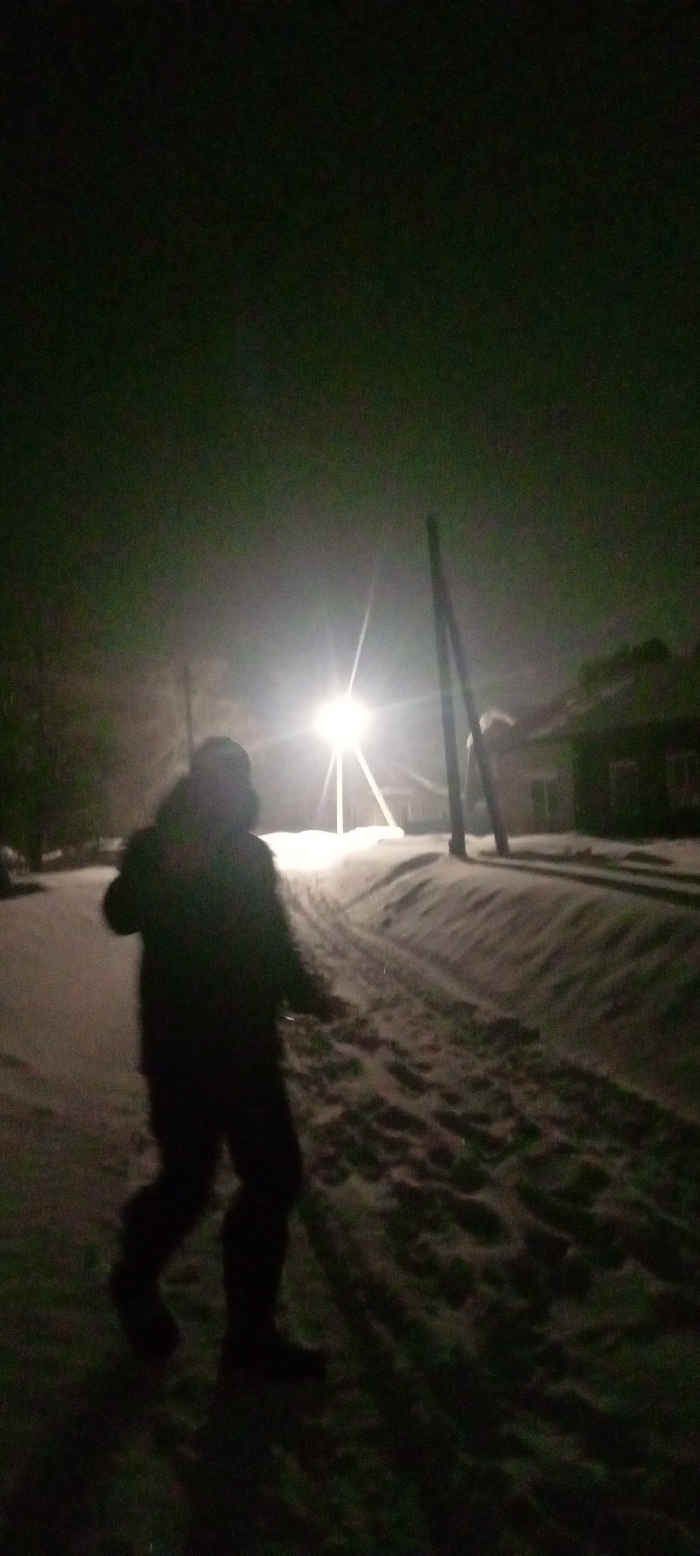 The image captures a nighttime, snow-covered street scene in Красный Маяк, Russia. In the foreground, a single individual is depicted in silhouette, walking towards the midground on a path heavily laden with snow. The person wears a bulky winter coat with a hood and dark lower garments. The snow on the path shows multiple depressions and tracks. A bright streetlight in the midground acts as the primary light source, creating significant glare and a starburst effect. Adjacent to the light, two dark utility poles ascend vertically. To the right and behind the streetlight, several dark, indistinct structures, consistent with residential buildings, are visible with snow partially covering their roofs. The sky is dark, and the environment suggests a winter setting with fresh or accumulated snowfall.