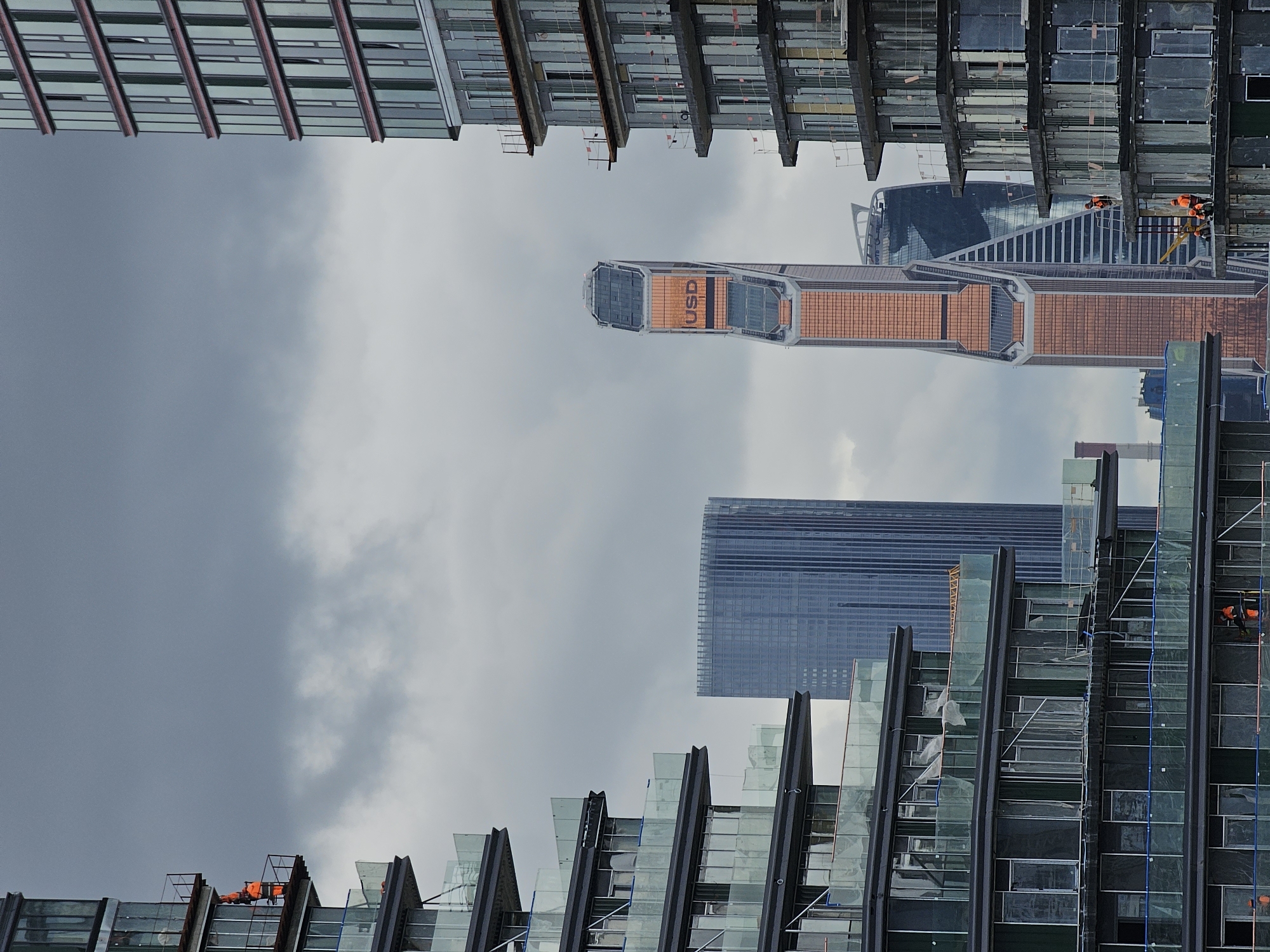 The image captures a segment of Moscow City under a cloudy sky. In the foreground, two residential buildings under construction are partially visible, one on the left and another on the right, showcasing their tiered, balcony-heavy designs and unfinished state. Several construction workers in bright orange vests are seen on the balconies of the building on the right, engaged in work. In the background, the iconic skyscrapers of Moscow City stand prominently: the East Tower of the Federation Tower, distinguished by its copper-colored facade and the inscription "USD" on its upper section, and the Naberezhnaya Tower to its left, recognizable by its dark glass and angled silhouette. A blue-toned, rectangular skyscraper is also visible behind the construction on the left. The overall scene depicts a dynamic urban landscape of ongoing development against a dramatic, overcast sky.