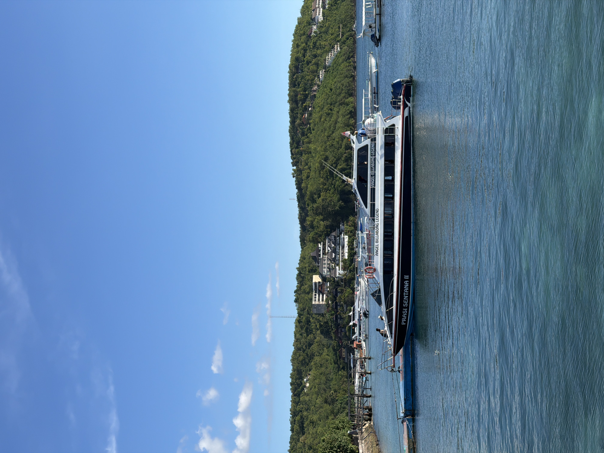 This image captures a vibrant coastal scene in Nusa Penida, Indonesia, under a clear blue sky. The main subject is a large, sleek white and dark-colored boat, possibly a fast ferry or tour vessel, docked in calm, clear blue-green waters. Its name, "PRASI SENTANA II", is prominently displayed on its side, along with "ANGEL BILLABONG LEGEND" partially visible higher up.
The boat appears to be at a pier or port, with a few small figures visible on its deck, suggesting passengers or crew. Behind the main vessel, other smaller boats are also docked. The water's surface shows subtle ripples, reflecting the bright daylight.
To the right of the water, a verdant, densely forested hill rises steeply, covered in lush green tropical vegetation. Scattered among the trees on the hillside are several white buildings, likely resorts or residences, blending into the natural landscape. A tall, slender antenna or tower is visible atop the distant part of the hill.
The sky above is a brilliant, unblemished blue, with only a few wispy white clouds adding texture, indicating excellent weather conditions. The overall impression is one of a sunny day at a busy yet picturesque island harbor, ready for transport or tourism activities. The strong natural light suggests it is midday or early afternoon.