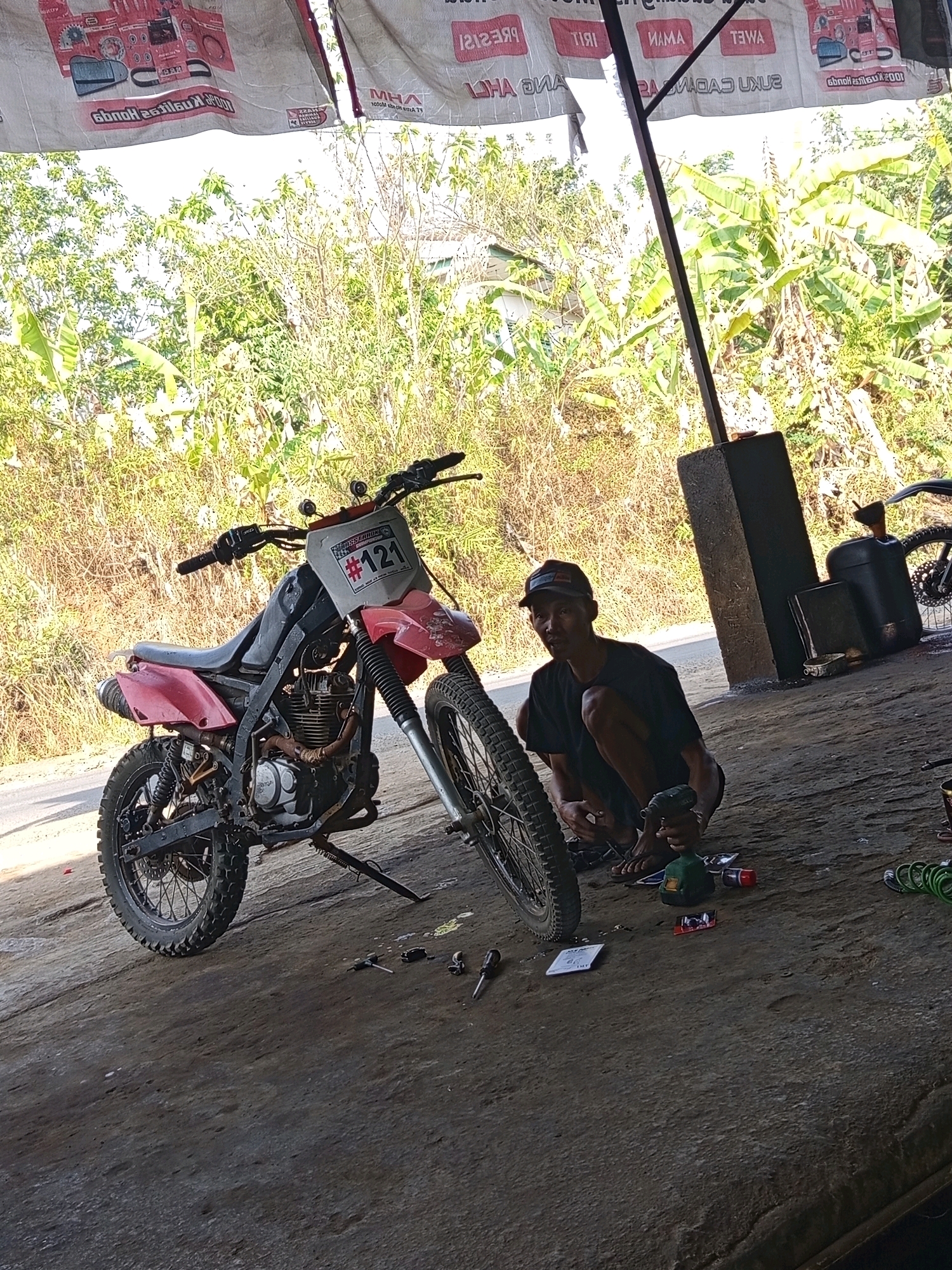 A man wearing a dark t-shirt and a baseball cap is crouched on a dirt floor next to a dirt bike. He is holding a green and black power tool, possibly a drill, in his right hand. The dirt bike is red and black with the number "#121" on a white plate on the front. The scene appears to be an outdoor workshop or repair area. Behind the man and the bike, there are banana trees and other foliage, suggesting a tropical or subtropical environment. A large black fuel can and a smaller black container are visible behind the man. The ground is covered in dirt and there are various tools scattered around the area. The image is taken from a slightly elevated angle.