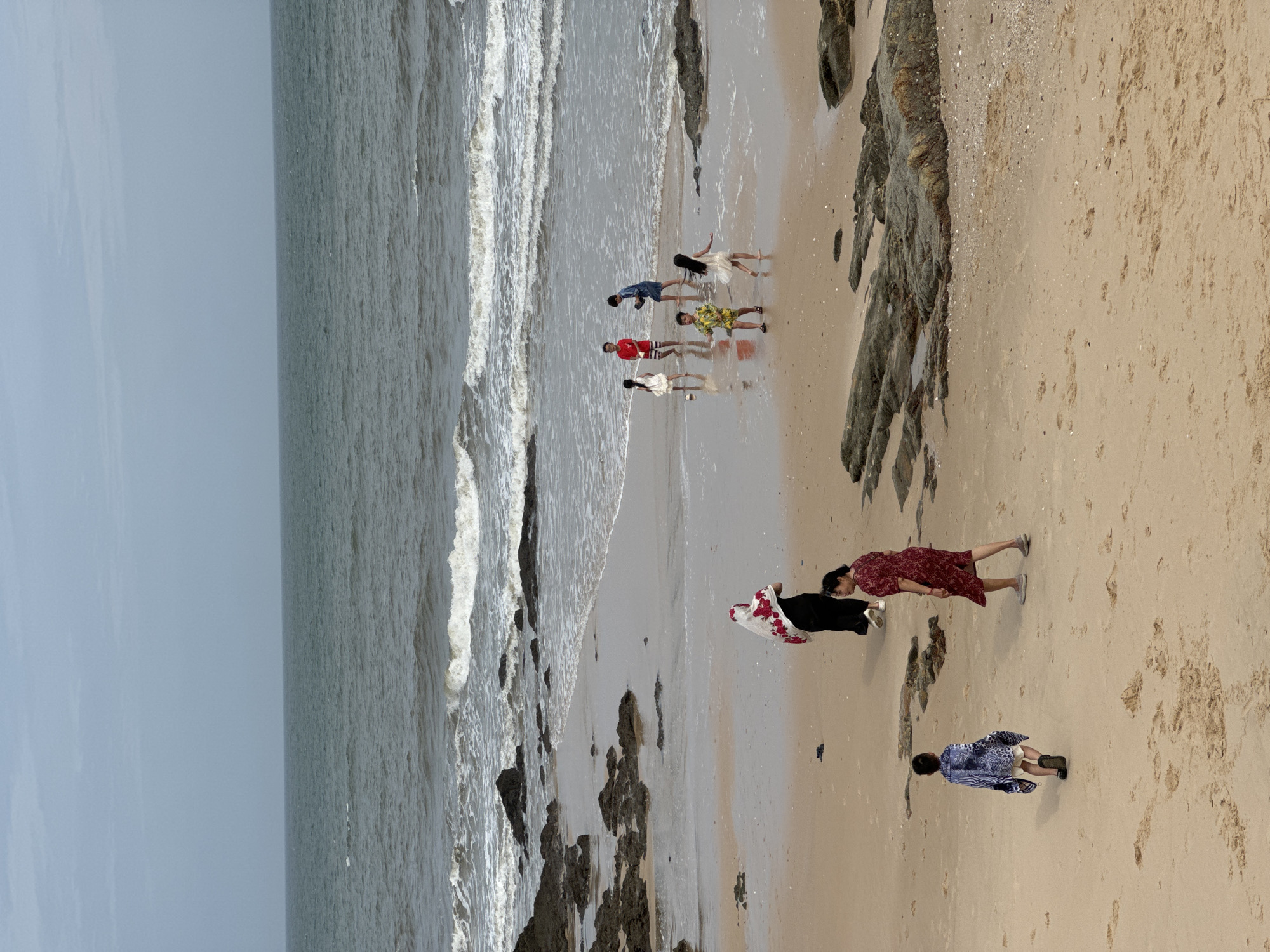 The image captures a serene beach scene in Mũi Né, Vietnam, under an overcast sky. The foreground features a sandy beach with scattered rocks and the gentle lapping of waves from the ocean. Several individuals are present on the beach.
In the mid-ground, a group of children is playing near the water's edge. One child, wearing a yellow and green patterned shirt, is in motion. Another child in a white dress stands to their right. Further back, a child in a red shirt and another in a white dress are also visible, along with a child in a blue denim outfit.
Closer to the foreground, two women are walking. One is wearing a black dress and a white headscarf with red floral embroidery, while the other is in a red patterned dress. A young child in a blue and white patterned jacket and white shorts walks away from the viewer towards the left. The ocean stretches out to the horizon, with waves creating white foam as they meet the shore. Rocks are visible along the coastline, some partially submerged in the water. The overall atmosphere is tranquil, with the natural beauty of the Mũi Né coastline being the primary focus.