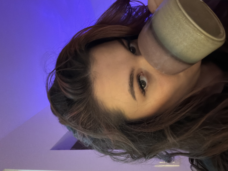 A person with brown hair drinks from a light green ceramic mug. Their eyes are visible, gazing towards the viewer. The background is split, with a purple-lit wall above and a brighter, reflective surface below. The camera angle is from below, looking up at the person and mug. The image is rotated to the side.