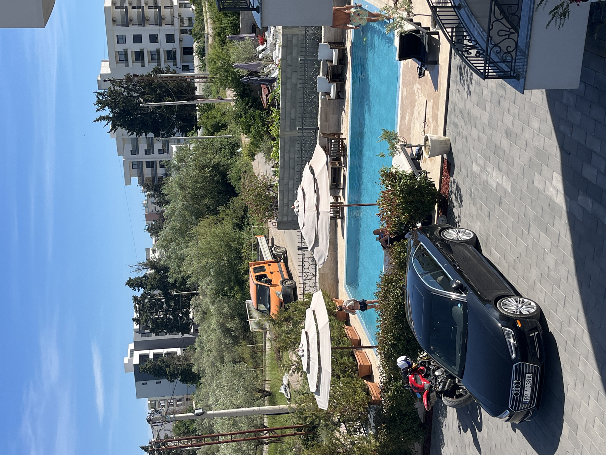 In a courtyard adjacent to a swimming pool, a black Audi sedan is parked on a paved area. To the left of the Audi, a red motorcycle is positioned. The swimming pool is surrounded by greenery and lounge chairs. Two individuals are visible near the pool, one standing and partially in the water, and another walking away from the camera. In the background, an orange truck is parked on a road, with several modern apartment buildings visible behind it. The sky is clear and blue. The scene appears to be in the city of Ulcinj - Ulqin, Montenegro.