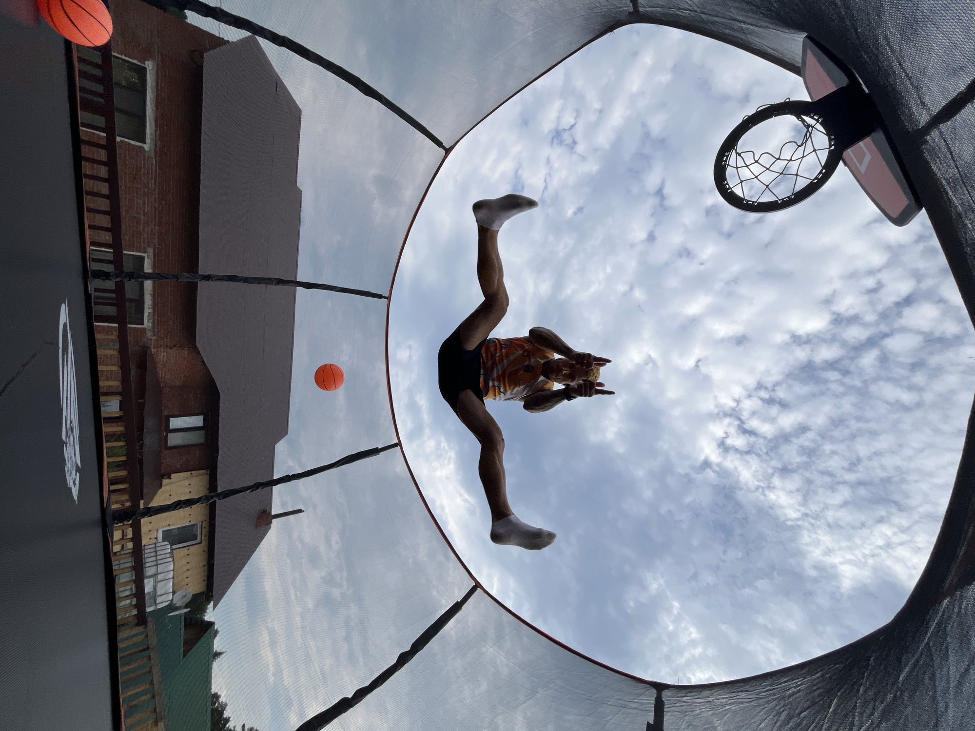 The image captures a low-angle, wide-angle shot from within a trampoline enclosure, looking upwards towards the sky and a person performing a split jump. The person, a light-skinned male with blond hair and tattoos, is airborne in the center of the frame, wearing an orange and white patterned sleeveless shirt, black shorts, and white socks. He is mid-jump, with legs extended wide apart in a split. His arms are raised, and he is making a "peace" sign with both hands, with his index and middle fingers extended upwards.
Above him, a basketball hoop is visible at the top left of the frame. In the background, a partially visible orange basketball is suspended in the air. Below the trampoline's net and the person, a residential area with brick and yellow-clad houses with dark brown and green roofs is depicted. A wooden fence is also visible in the foreground. The sky is partly cloudy, with scattered white clouds against a pale blue background.
The image appears to be taken outdoors, likely in a backyard, given the presence of a trampoline, house, and fence. The location is specified as Chaikovsky, Russia. The overall scene suggests a moment of recreational activity, possibly during daytime given the sky conditions.