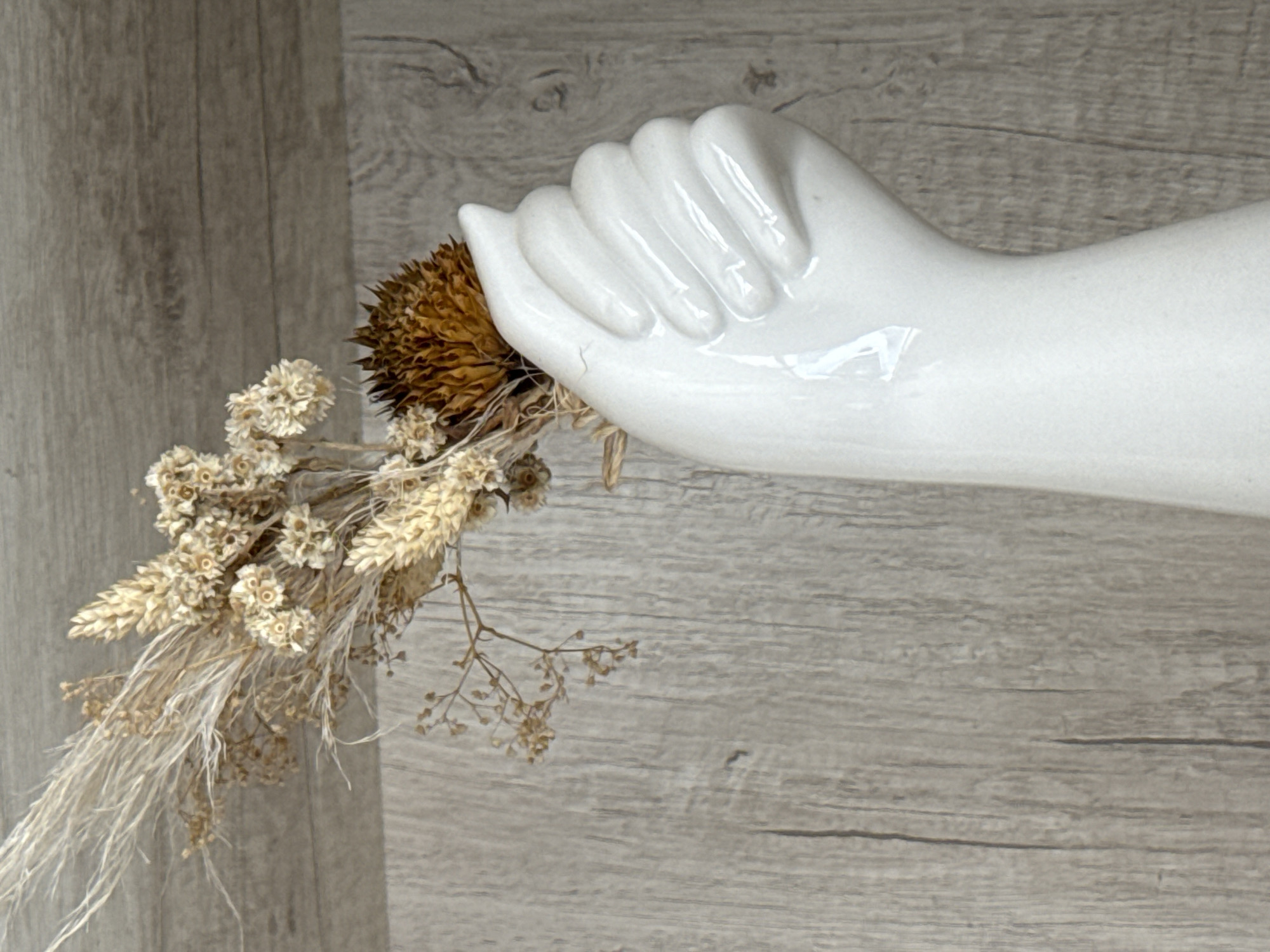 A still life photograph captures a white, glossy ceramic hand sculpture holding a bouquet of dried flowers and grasses. The ceramic hand is positioned on the right side of the frame, extending horizontally with its fingers gently curled, as if cradling the floral arrangement. The bouquet consists of a large, spiky, dark brown dried flower, clusters of small, light beige papery flowers, and thin, wispy dried grasses and stems that cascade downwards and to the left. The background is a light-colored wooden surface with visible grain patterns, creating a rustic and textured backdrop. The image features a blend of smooth, artificial elements (the ceramic hand) and natural, organic textures (the dried botanicals and wood). There is no discernible activity, text, specific time of day, or weather information within the frame. The overall aesthetic is one of natural decor and muted, earthy tones.