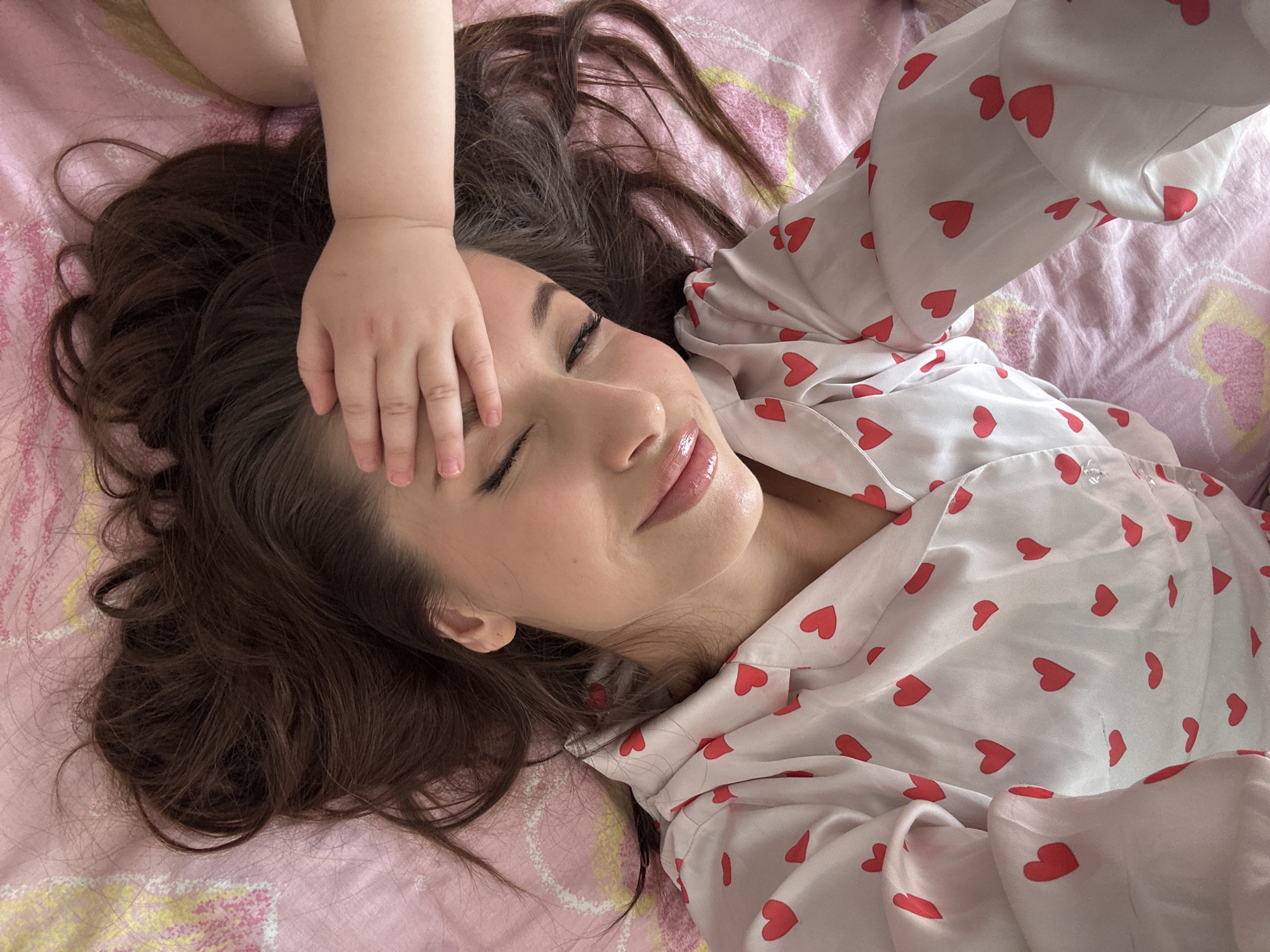 A woman is lying on a pink bed with a patterned sheet, wearing a white shirt with red heart print. A child's hand rests on the woman's forehead. The location is unknown and is not evident in the image.