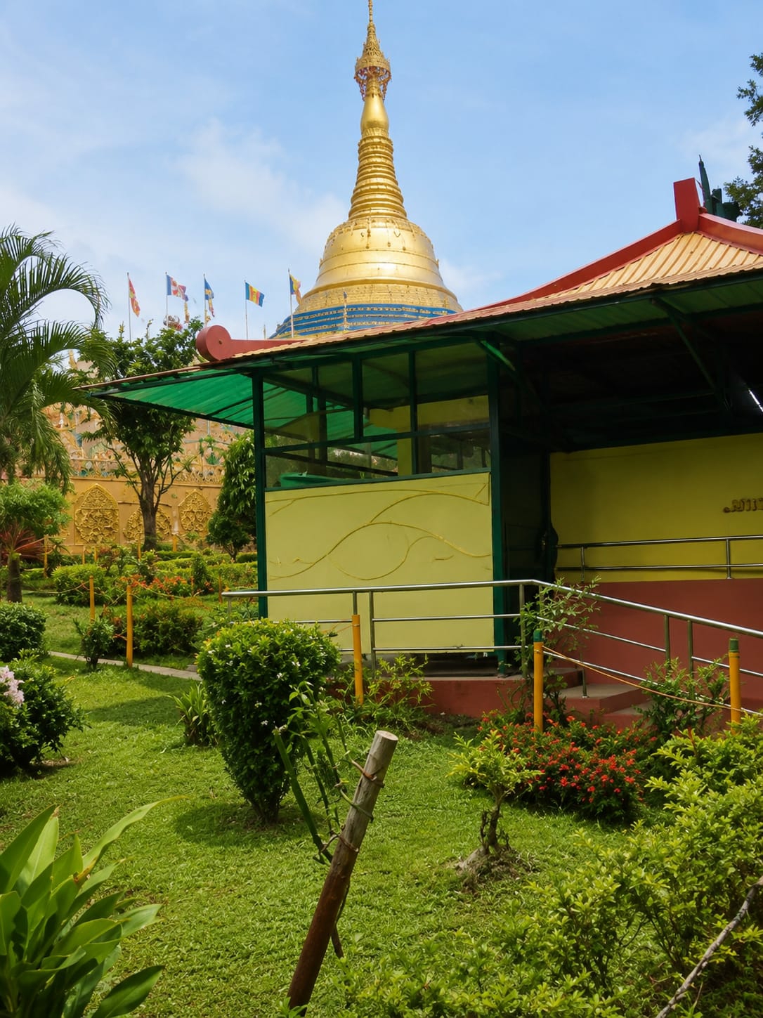 The image shows a golden Buddhist stupa with a tiered roof, situated behind a covered walkway with a green translucent roof supported by green metal poles. The stupa is adorned with blue bands and intricate gold detailing. In front of the stupa, a yellow wall with a wavy pattern is visible, with a metal railing along the walkway. The area is landscaped with lush green grass, manicured bushes, and flowering plants in shades of red and pink. Several flags are displayed on poles in the background. The sky is clear blue with scattered white clouds. The location is identified as the city of Dolat Rayat, Indonesia.