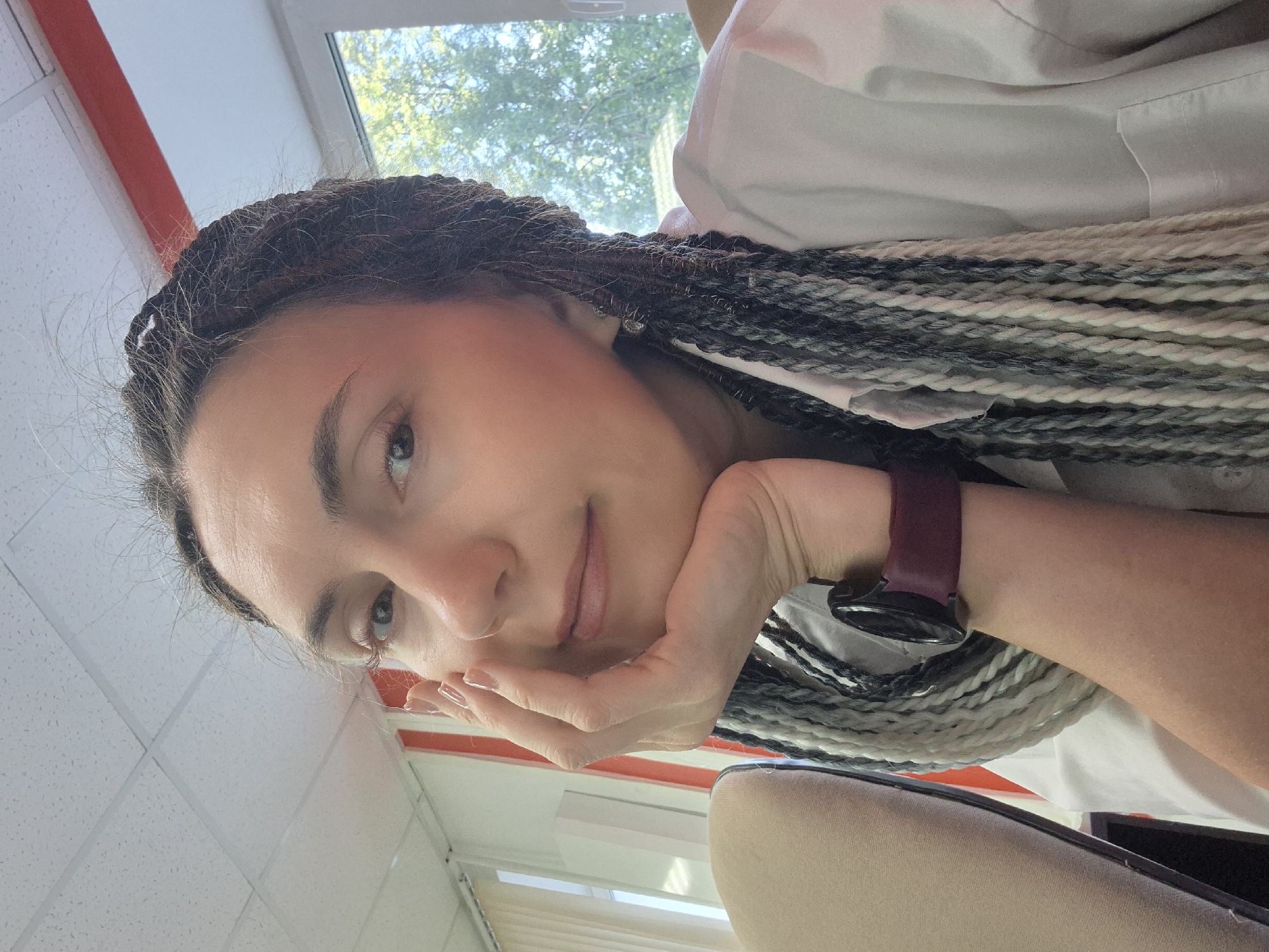 A close-up, waist-up selfie is presented of a Caucasian woman with braided hair, appearing to be indoors, possibly in an office. Her hair is styled in numerous thin braids, a mix of dark brown, grey, and white. She is wearing a white collared shirt. Her right hand is resting on her chin, and she is looking directly at the camera with a neutral expression. A smartwatch with a dark red band is visible on her left wrist. The background includes a window with trees and greenery visible outside, suggesting daylight. The interior features a white tiled ceiling and what appears to be office furniture or partitions. The image lacks specific identifying markers for the city of Chaikovsky, Russia, and provides no details about the woman's activities or interactions beyond posing for the selfie.