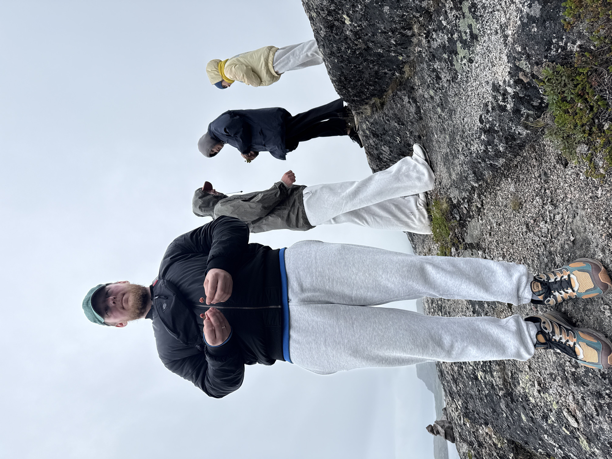 The image depicts a group of five individuals on a rocky, overcast terrain, likely near the coast. In the foreground, a man wearing a green cap, black jacket, and light gray sweatpants holds two small, reddish objects in his hands. He is also wearing a black fanny pack and colorful athletic shoes. Behind him and to his right, four other people are visible, all wearing hooded jackets and looking downwards. One person in a dark jacket and black pants appears to be examining something in their hands. Another, wearing a yellow jacket, stands slightly apart. The landscape features dark, rugged rocks with patches of sparse green vegetation. In the distance, a body of water and a hazy coastline with rock formations are discernible. The overall atmosphere is muted due to the cloudy sky. The location is stated to be Teriberka, Russia, suggesting a subarctic coastal environment.
