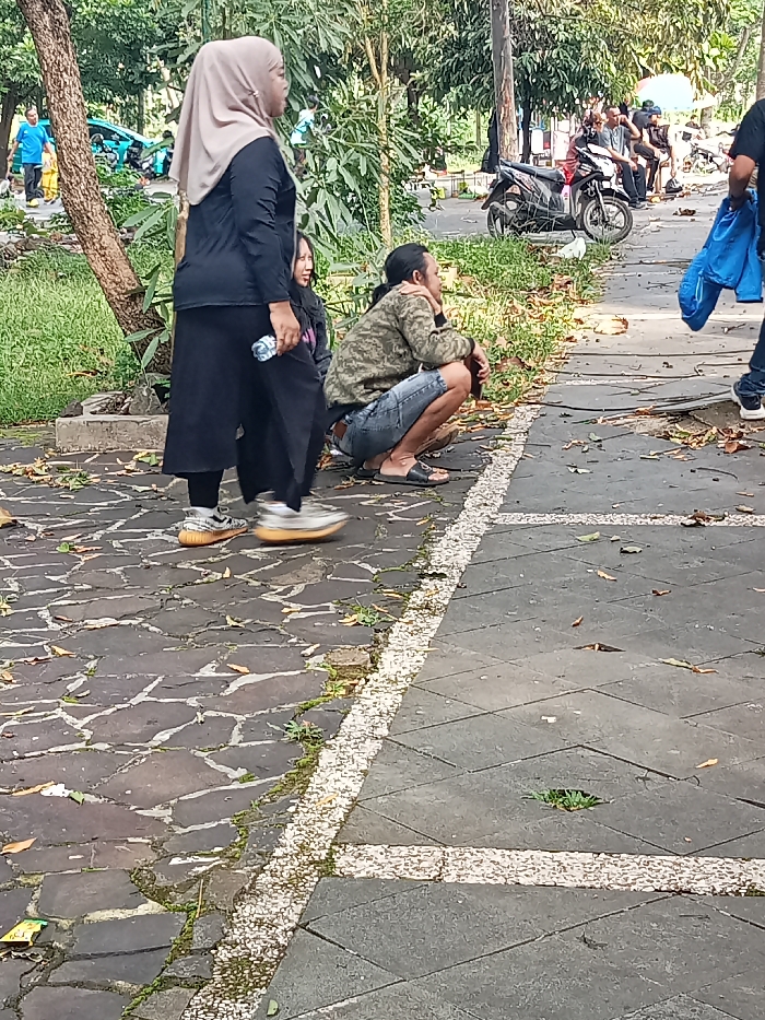 The image captures an outdoor scene in Singaparna, Indonesia, likely a public park or pathway, during daylight hours. In the mid-ground, a woman wearing a light brown hijab, a black long-sleeved shirt, and a long black skirt or pants is walking towards the right side of the frame. She is holding a plastic water bottle in her right hand, and her left foot, clad in light-colored sneakers with prominent orange soles, is blurred, suggesting motion.
Behind her, partially obscured, is another person. To her right, a man with long hair tied back is squatting or crouching by the edge of the path. He wears a camouflage-patterned long-sleeved shirt, denim shorts, and black sandals. His head is turned slightly to the right, and his left hand rests on his chin. On the far right, a person in a blue jacket is partially visible, also appearing to be in motion.
In the background, several more people are gathered or resting, some sitting. A dark-colored scooter or motorbike is parked further down the path. The area features a mix of lush green foliage, trees, and grass. The ground consists of two types of paving: irregular, dark grey or black flagstones on the left, and lighter grey square tiles with a distinctive white pebbled border on the right. The overall atmosphere appears relaxed, with people engaged in casual outdoor activities. No discernible text is visible in the image.