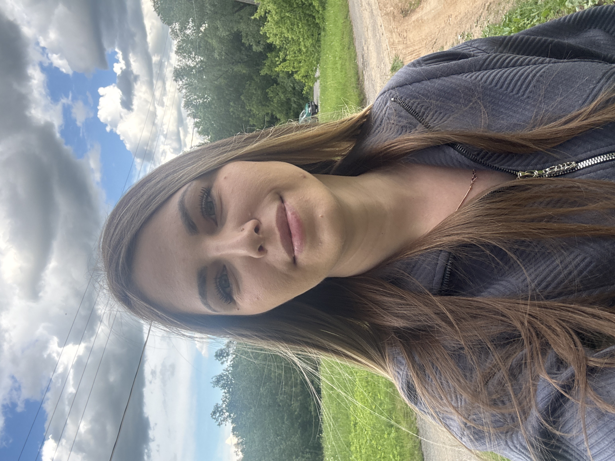 A woman is posing for a selfie outdoors. She has long brown hair and is wearing a gray jacket. In the background, there's a partly cloudy sky, trees, and a road. The location is a rural or suburban area, possibly near Shevlyakovo, Russia. Power lines are visible in the sky. The lighting suggests it's daytime.