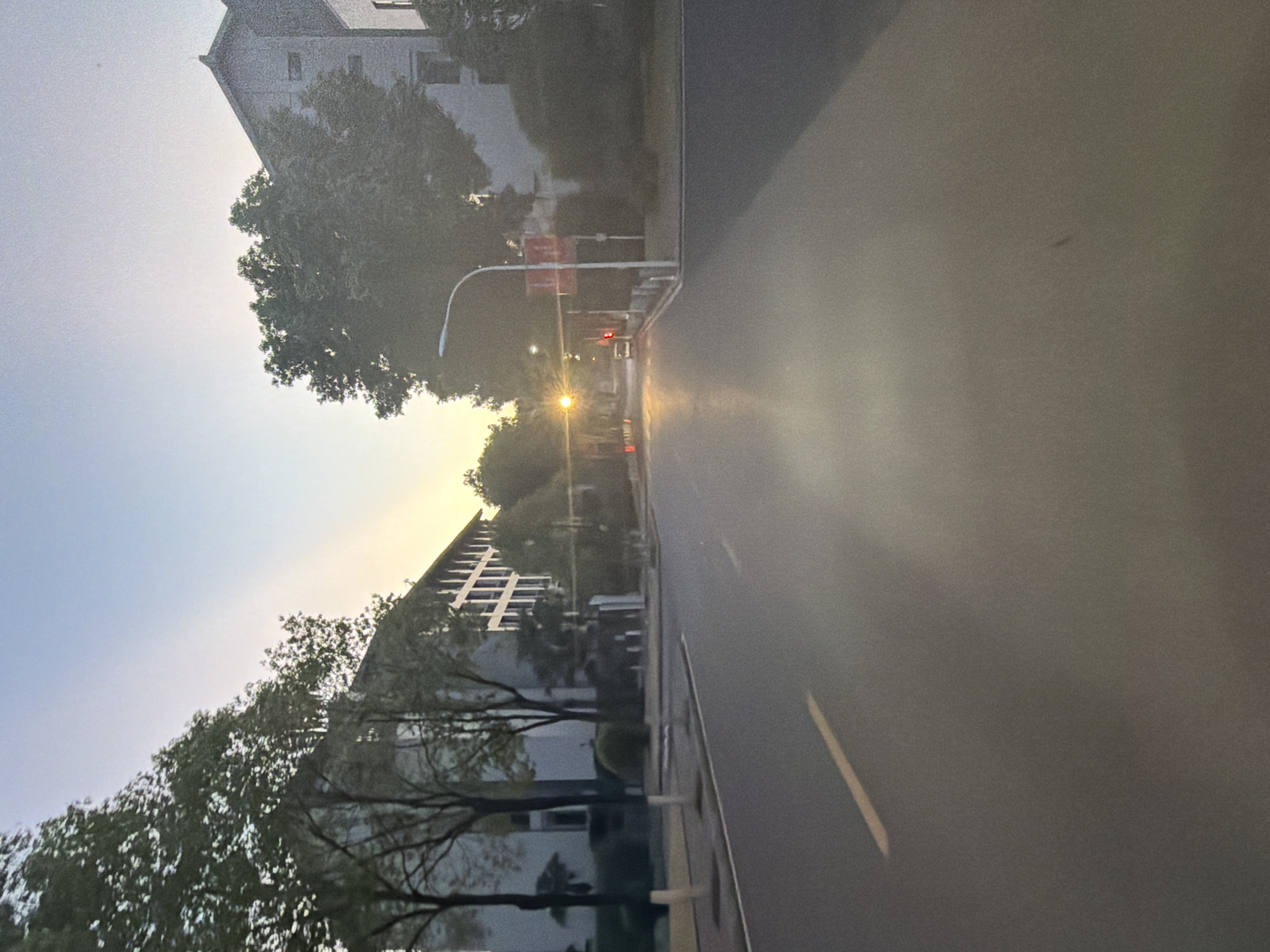 The image captures a street scene in Jiangjin District, Chongqing, China, during what appears to be sunrise or sunset. A paved road dominates the foreground, with a yellow line marking the lane. To the left and right of the road are buildings with multiple windows, flanked by trees and some shrubbery. A street light is visible on the right side of the road, and further down the street, a traffic light shows a red signal. The sky is a gradient of blue and pale yellow, indicating the low angle of the sun. The overall impression is one of a quiet urban environment at dusk or dawn.