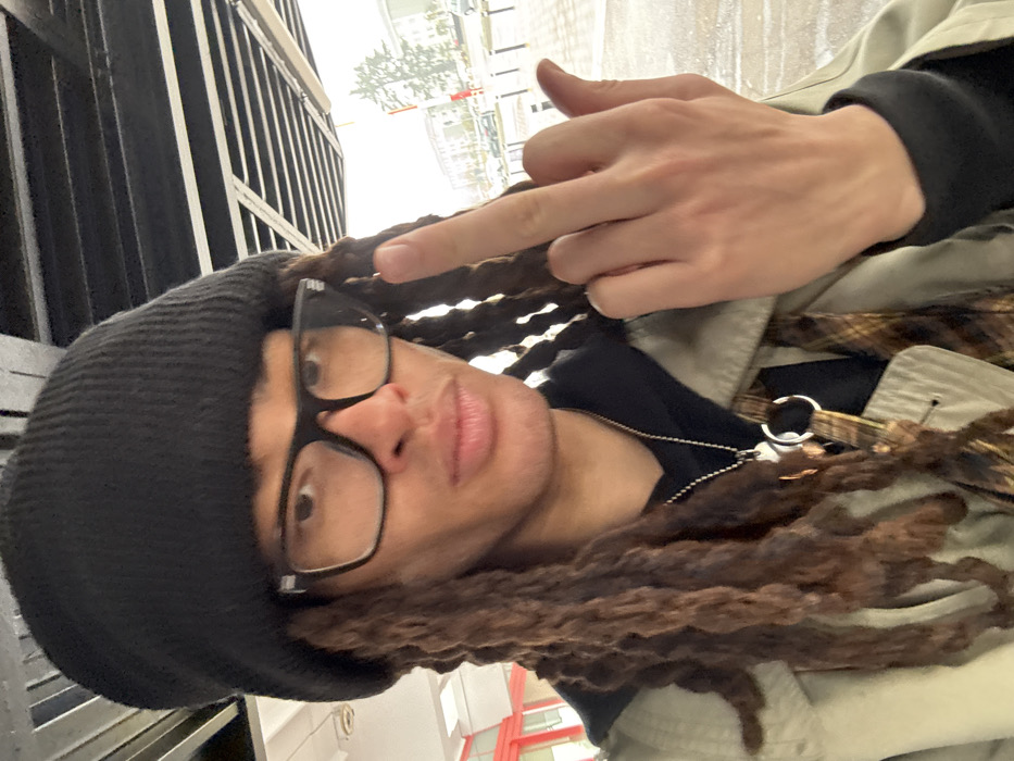 A light-skinned individual with brown dreadlocks, wearing a black knit beanie and dark-rimmed glasses, is depicted from the chest up, outdoors in Somma Lombardo, Italy. The individual wears a black hooded garment, a light beige trench coat, and a brown plaid garment visible underneath. A silver-toned ball chain necklace with a pendant and a ring is present around the neck. The subject's right hand is raised, with the middle finger extended. The background reveals an overcast sky, a wet paved surface indicating precipitation, multiple parked vehicles, and multi-story buildings. A dark, slatted overhead structure is visible above the subject. A red and white striped pole is present in the distant background to the right, and a red structural element is partially visible on the left.