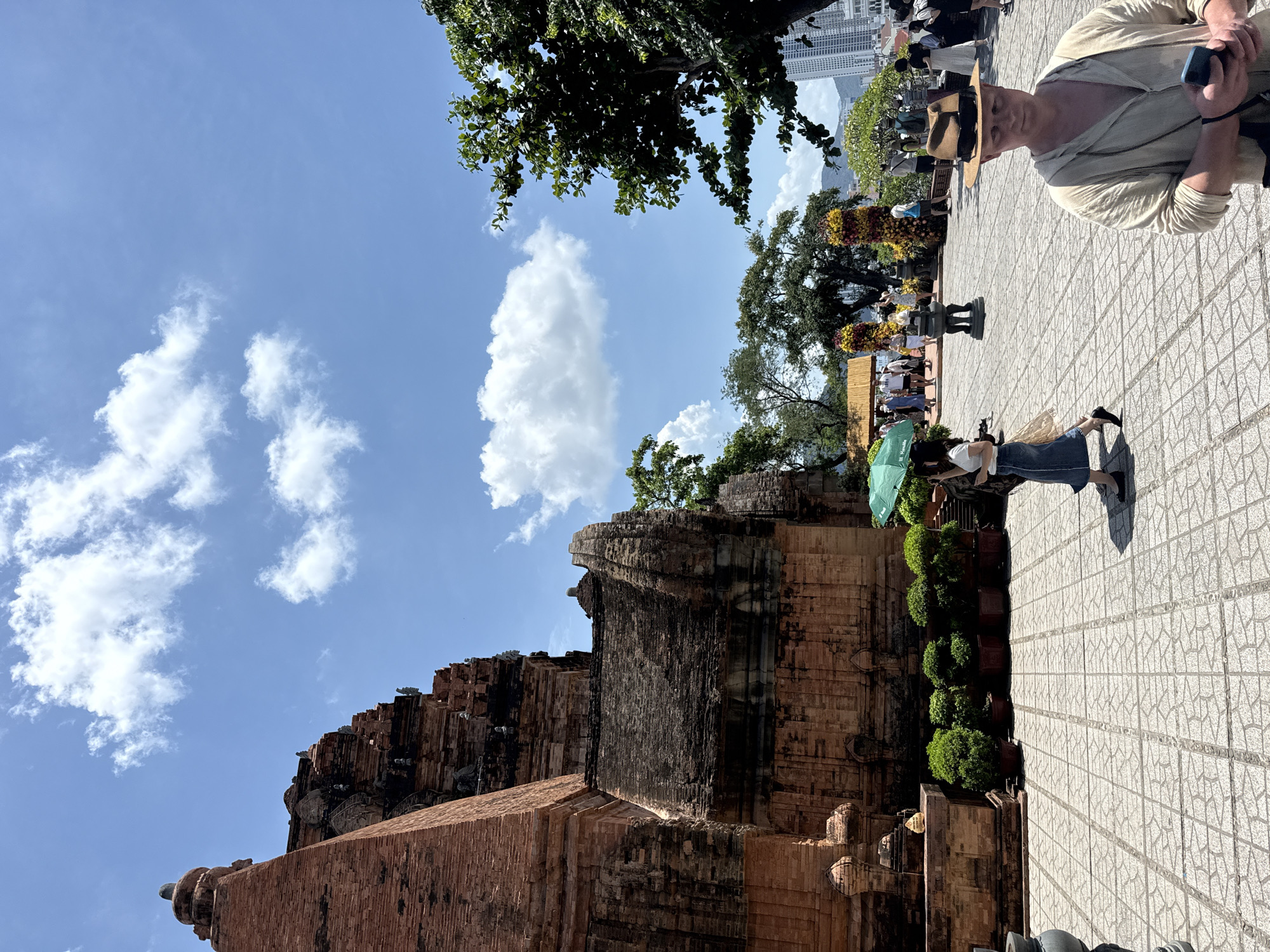 This photograph shows a daytime view of the Po Nagar Cham Towers in Bac Nha Trang Ward, Vietnam. The brick structures of the towers occupy the left side of the frame, reaching towards a blue sky with scattered clouds. To the right, a paved area is visible with several people. One person is holding a phone and wearing a straw hat. Another person stands with a green umbrella. There are potted plants along the border of the towers and paved area. In the background, there are trees and buildings including high-rise apartments.