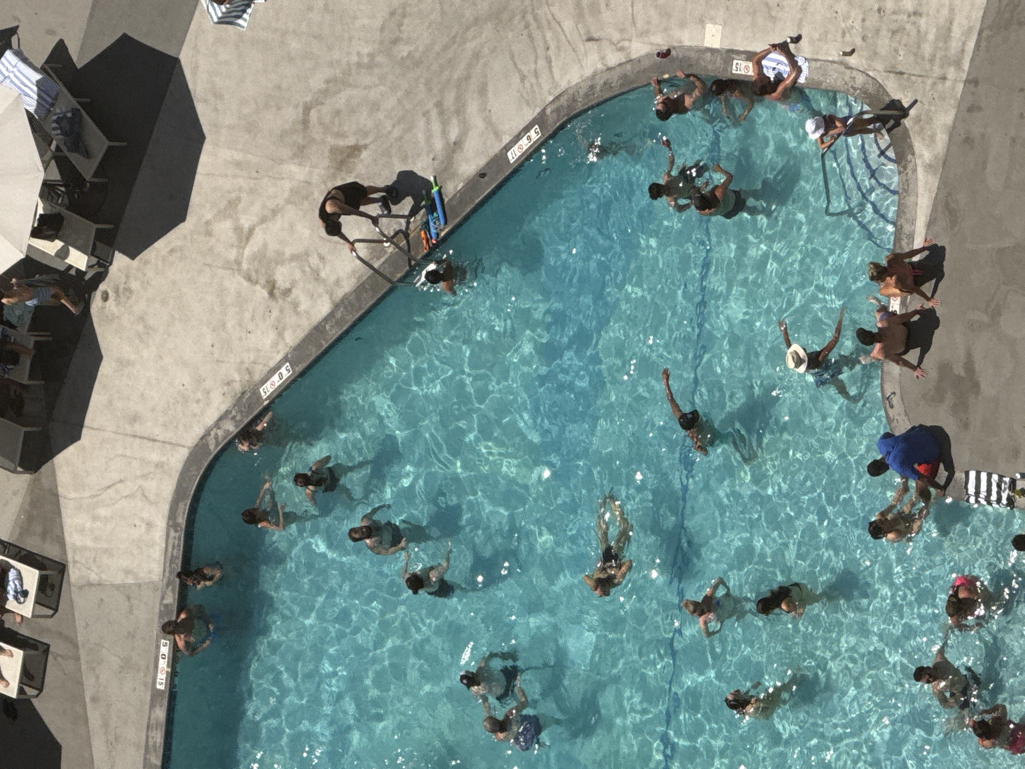 An aerial view captures a crowded, sunny outdoor swimming pool with a concrete deck. Numerous people are enjoying the water, with many swimming, splashing, and interacting. Some individuals are standing at the edge of the pool, while others are relaxing on lounge chairs under umbrellas on the deck. The pool has depth markers indicating 5 and 15 feet. The scene suggests a recreational gathering at a hotel or public pool. The specific location in Chicago, United States of America, is not discernable from the image.