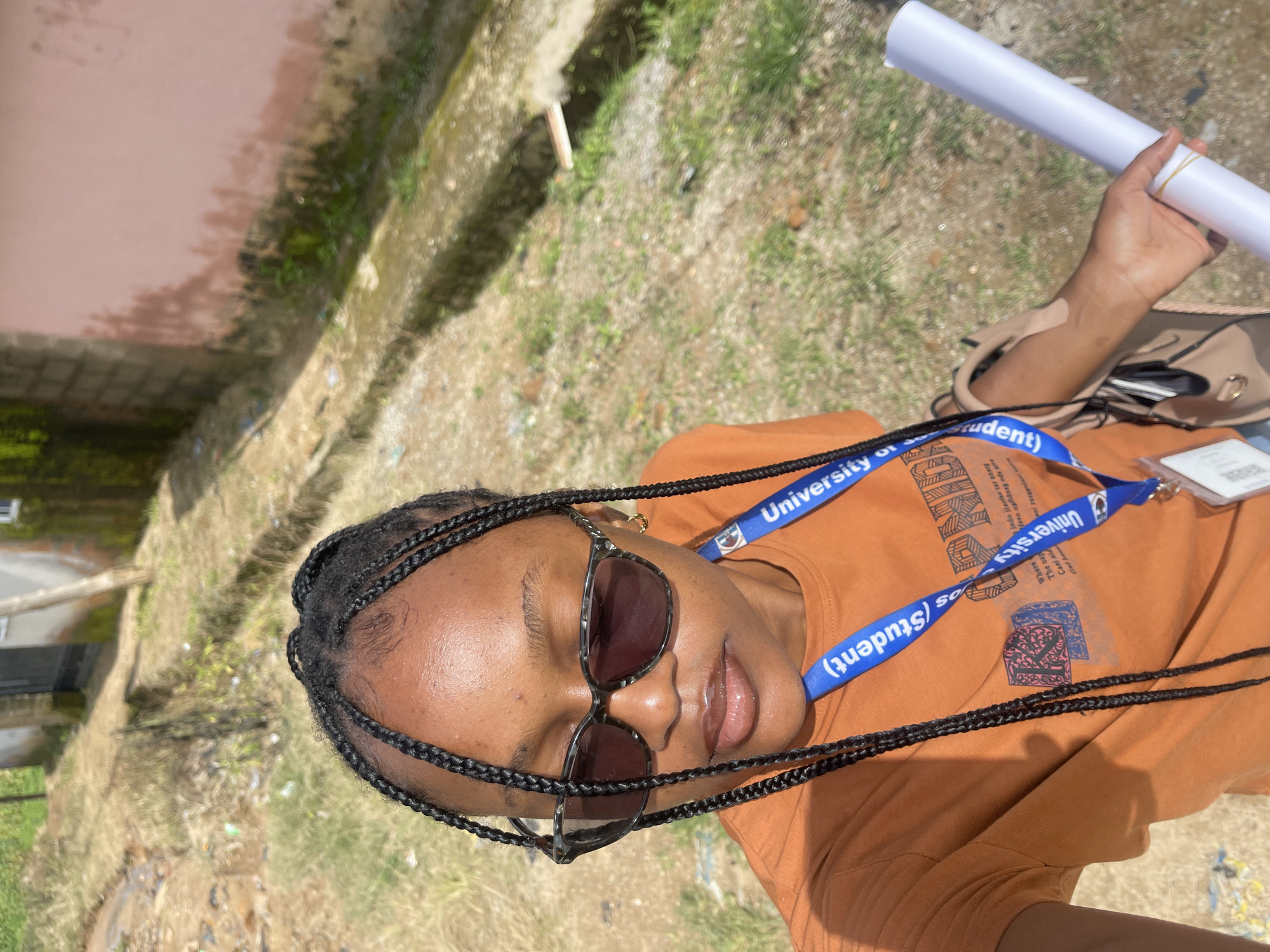 In the city of Jos, Nigeria, a young Black woman with braided hair and sunglasses takes a selfie outdoors. She wears an orange t-shirt with a blue lanyard around her neck, which displays "University of... (Student)". In her right hand, she holds a rolled-up white paper. The background shows a dirt path with some overgrown grass, a drainage ditch, and parts of buildings, one with a pink wall.