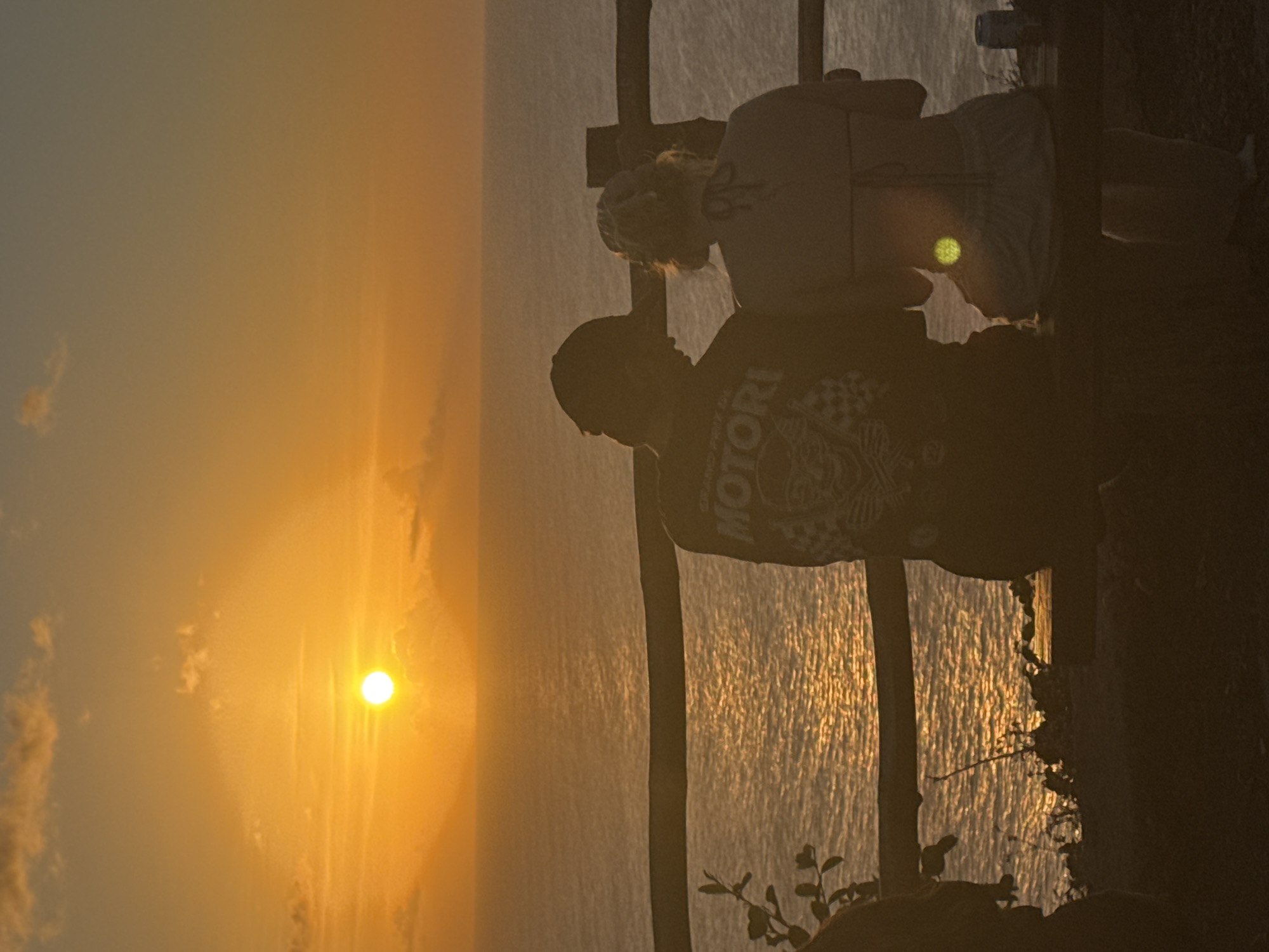 An outdoor, eye-level, medium shot captures a silhouetted couple seated on a wooden bench, facing the ocean at sunset. The man, on the left, wears a baseball cap and a t-shirt with a graphic design featuring the word "MOTORI". The woman, on the right, has her hair in a bun and wears a bikini top and light-colored pants. Both are positioned in the foreground, with their backs to the viewer. Between them and the water is a wooden fence. The ocean is visible in the midground, reflecting the warm orange and yellow hues of the setting sun. The sky is a gradient of orange and yellow, with scattered clouds. The scene is set against a backdrop of the ocean and the sunset.