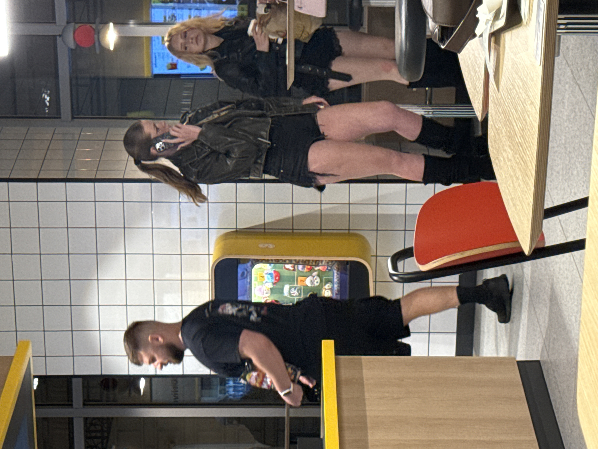 In a brightly lit, modern indoor setting with white tiled walls and light-colored flooring, a man with a beard and short hair, wearing a black t-shirt and black shorts, is standing near a counter. He appears to be looking towards a yellow gaming machine mounted on the wall, which displays colorful graphics. To his right, a woman with long brown hair tied in a ponytail, wearing a dark leather jacket and black shorts, is holding a phone to her ear. Further to the right, another woman with blonde hair, wearing a black leather jacket and black shorts, is holding a paper cup and looking to her left. Tables and chairs are visible in the foreground and background, with one red chair prominently featured. The overall impression is that of a casual dining or entertainment establishment. The location is identified as Bratislava, Slovakia.