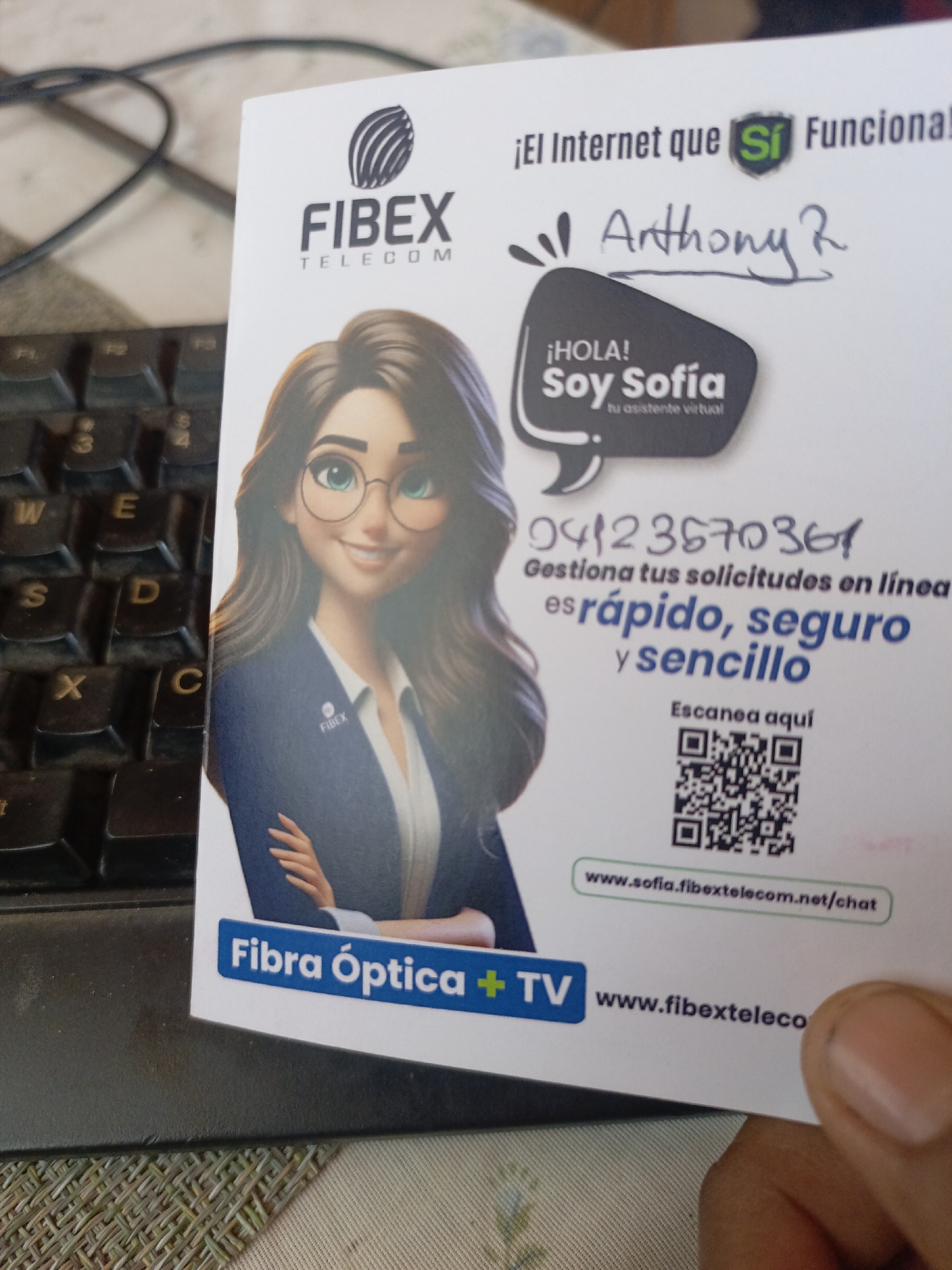 The image shows a business card for FIBEX Telecom, an internet provider, lying on a surface beside a keyboard. The card features a cartoon image of a woman with glasses, presumably representing "Sofia," a virtual assistant. The card also lists a phone number (04123570361), a QR code for scanning, and website addresses (www.sofia.fibextelecom.net/chat and www.fibexteleco). In handwriting, the name "Anthony R" is written on the card. "Fibra Optica + TV" is visible on the card. There's no visual evidence of the location of Juan Griego, Venezuela in the image.