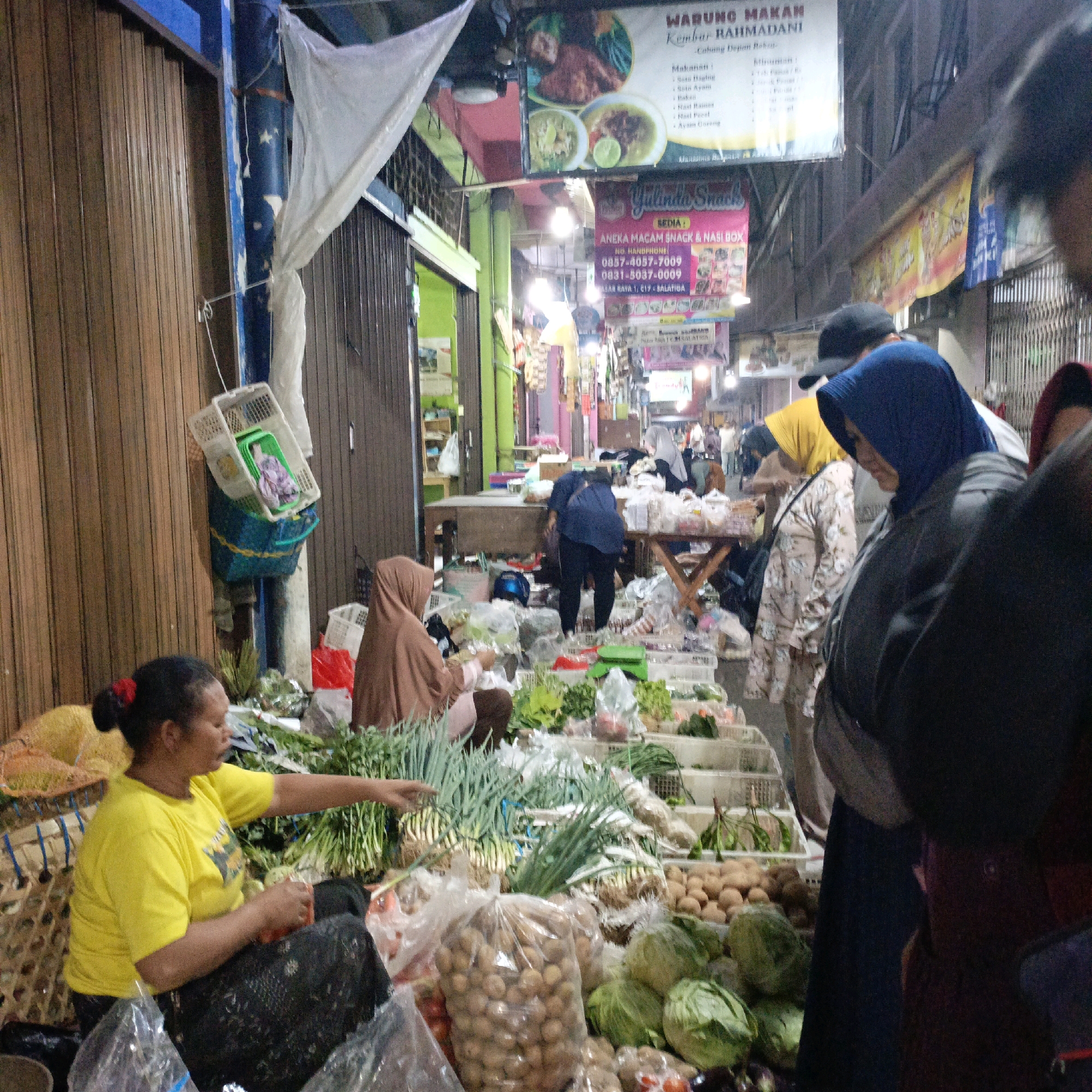The image depicts a street market in Salatiga, Indonesia. The scene is bustling with activity, featuring several vendors and customers amidst displays of fresh produce. In the foreground, a woman wearing a bright yellow t-shirt and a patterned dark skirt is seated and actively sorting vegetables, likely green onions, as she gestures with her right hand. Beside her, a large woven basket overflows with produce, and various plastic bags filled with potatoes and other vegetables are visible.
Behind her, a woman in a brown hijab is also seated, seemingly attending to her stall. Further into the market, other individuals are browsing or working. One person in a dark blue shirt and black pants is bent over a stall, and another person in a yellow hijab is visible in the background. Several market stalls are set up, with tables laden with a variety of vegetables like cabbage, potatoes, and green onions, all arranged in baskets and plastic bags. Signs are visible in the background, including one that reads "WARUNG MAKAN Kembar Rahmadani" and another for "Yulinda Snack". The environment suggests a lively, open-air market setting, possibly in a narrow street, with stalls extending down a walkway. The lighting appears to be a mix of natural light and artificial lighting from stall lamps.