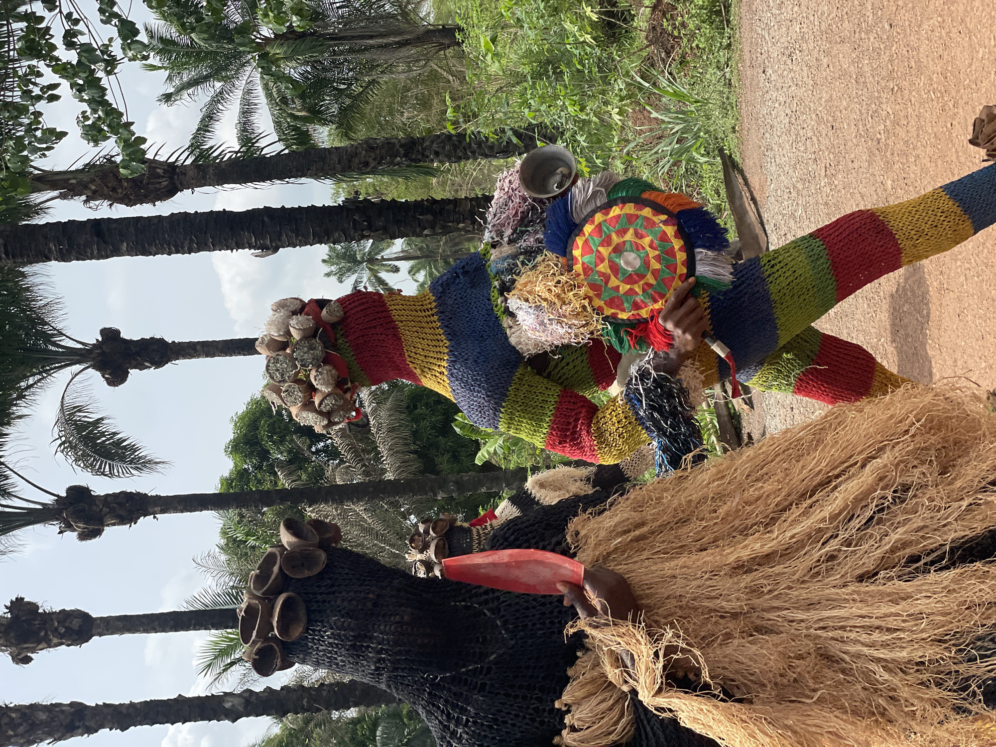 In Agba Umana, Nigeria, two masquerades stand outdoors on a sunny day with palm trees visible. One masquerade wears a black net-like mask adorned with small, round objects, possibly shells or nuts. It holds a red, elongated object. The other masquerade is more colorful, featuring a striped, knitted body, a circular headdress with a patterned design, and various hanging materials.