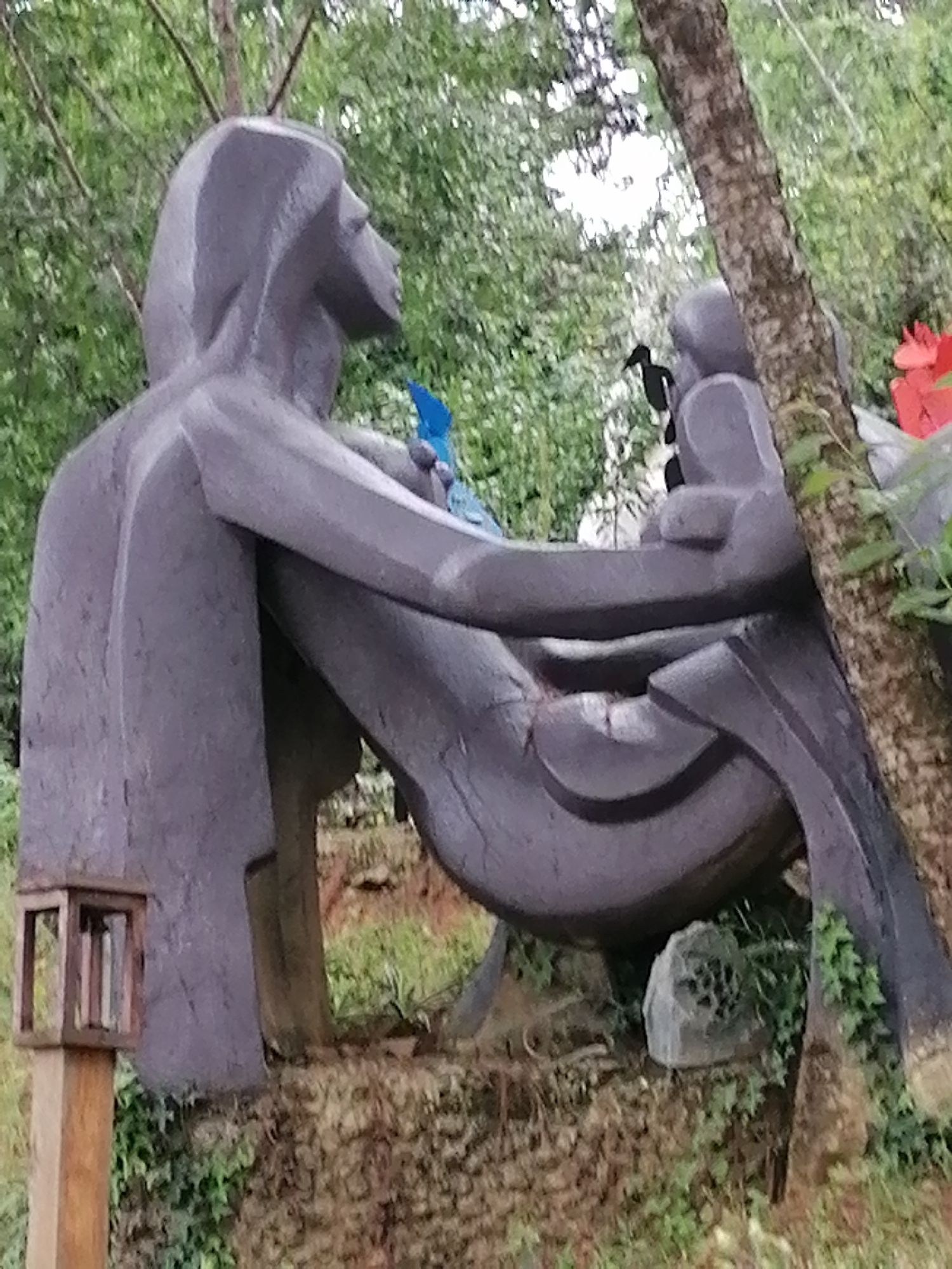 The image depicts a large, dark grey sculpture of two figures sitting together, possibly in a park or garden setting in Da Lat, Vietnam. The sculpture is made of what appears to be stone or concrete and has a stylized, abstract design. The figure on the left is larger and has its back to the viewer, with its head tilted to the right. The figure on the right is smaller and appears to be interacting with the larger figure, possibly holding a small blue object, which could be a bird. The sculpture is positioned on a mound of earth and moss, with vegetation and trees in the background. A small, wooden lantern-like object is visible on the left side of the image, attached to a wooden post. A tree trunk with rough bark is present to the right of the sculpture, obscuring some of the background. Red, flower-like objects are visible behind the sculpture on the far right. The overall scene suggests an outdoor art installation in a natural environment.