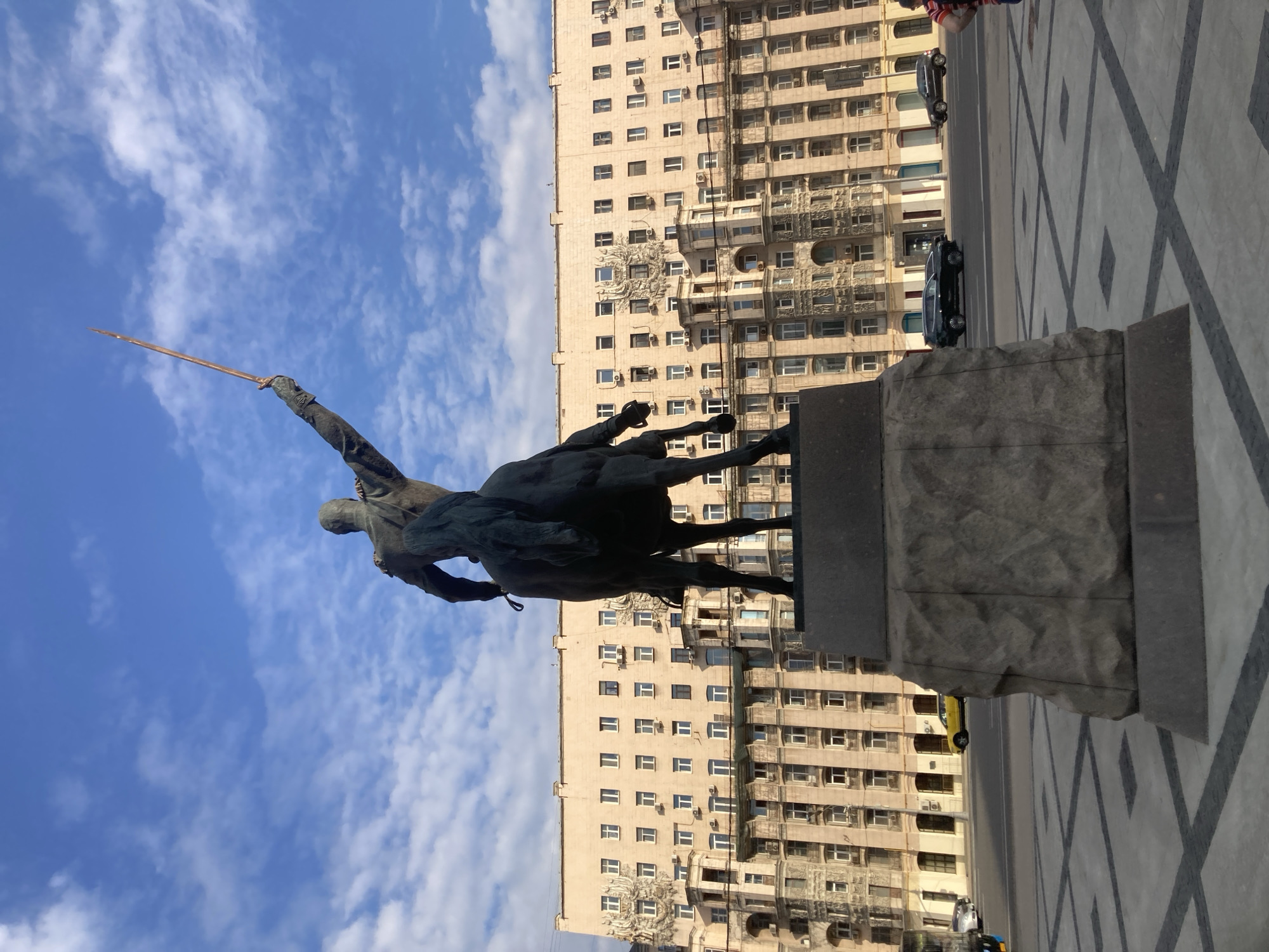 A bronze equestrian statue stands in the foreground, facing away from the camera. The rider, seen from behind, raises a spear upwards. The horse is depicted in mid-stride. The statue is mounted on a large, rough-hewn stone pedestal. Behind the statue, a massive, multi-story building dominates the background. The building has a light beige facade and features numerous windows, balconies, and ornate architectural details. The sky above is blue with scattered white clouds. In the middle ground, a street is visible with cars driving on it. A yellow car is particularly noticeable. The ground in front of the statue is paved with a geometric pattern of dark and light grey tiles. The scene appears to be in an urban setting, likely a public square in Moscow, Russia, given the architectural style of the building, which is characteristic of Stalinist-era architecture.