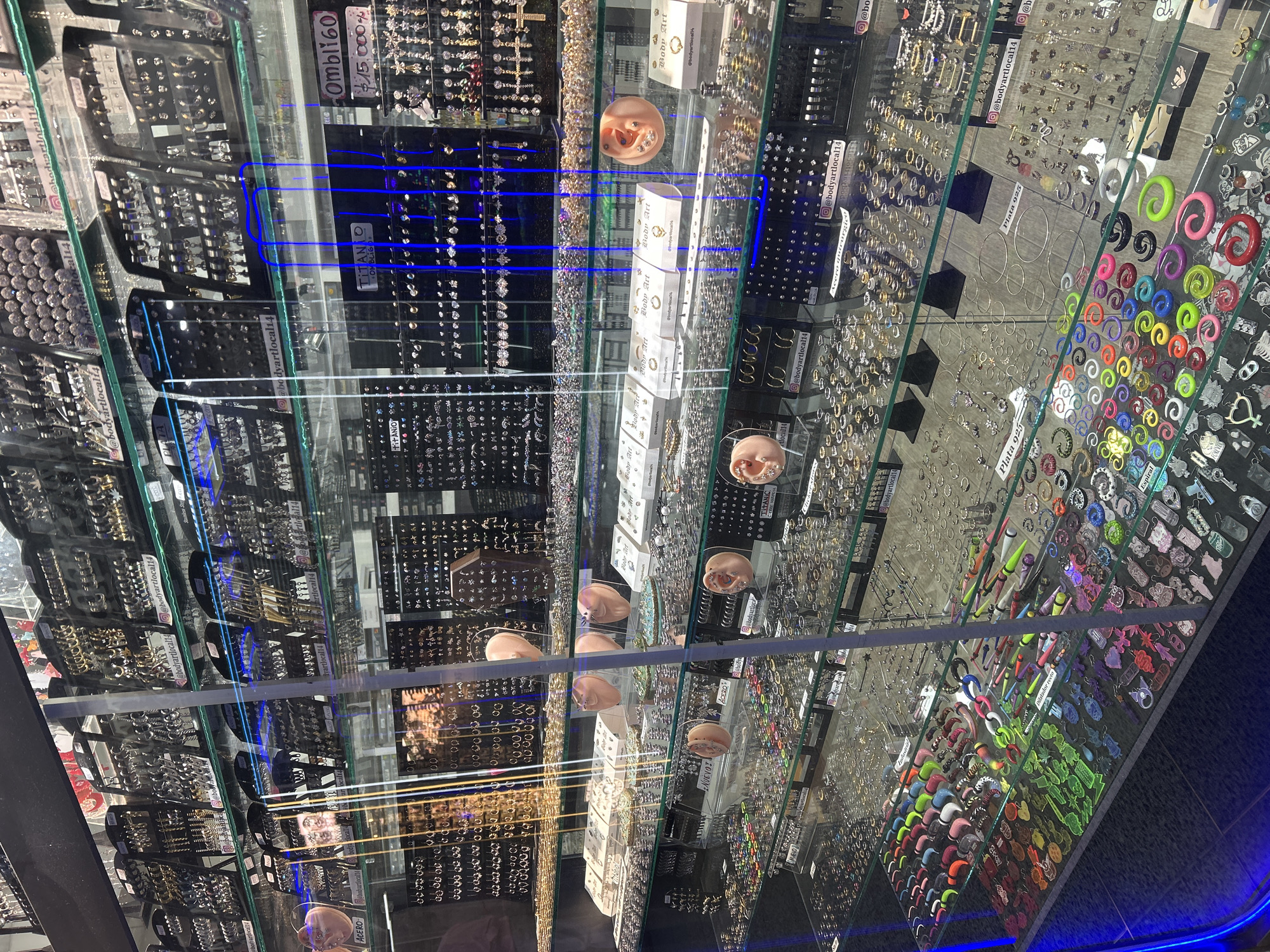 The image displays a bustling retail environment, specifically showcasing a wide array of body jewelry and piercings within illuminated glass display cases. The scene is viewed from an elevated angle, looking down into multiple shelves and compartments brimming with merchandise.
The primary subjects are the countless pieces of body jewelry, including various earrings, studs, hoops, rings, gauges, and spirals, meticulously arranged on black and white display stands. These items come in diverse materials and styles, ranging from delicate, crystal-adorned pieces to more robust, colorful gauges and plugs. Several flesh-toned artificial ear models are strategically placed throughout the displays, showcasing how different earrings and piercings appear when worn.
The activity depicted is the organized presentation of merchandise for sale in a specialized store, likely a body piercing studio or a unique jewelry shop in Buenos Aires, Argentina. The glass cases reflect blue neon lighting, creating a distinct ambiance and visual effect that sometimes causes glare on the reflective surfaces.
Notable details include the sheer volume and variety of the jewelry, catering to a wide range of tastes in body modification. Visible text on labels and small boxes identifies materials such as "TITANIO" (Titanium), "Acero" (Steel), "Acero quirúrgico" (Surgical Steel), and "Plata 925" (Sterling Silver 925). Specific product categories like "OMBLIGO" (belly button rings) and "Espiral" (spiral gauges) are also labeled. Social media handles like "@bodyartlocal14" are frequently visible, indicating the store's branding. A price tag of "$15,000" is discernible, likely in Argentine Pesos. The word "NUEVO!" (New!) highlights new arrivals. The overall impression is one of a comprehensive and well-stocked establishment for body art enthusiasts.