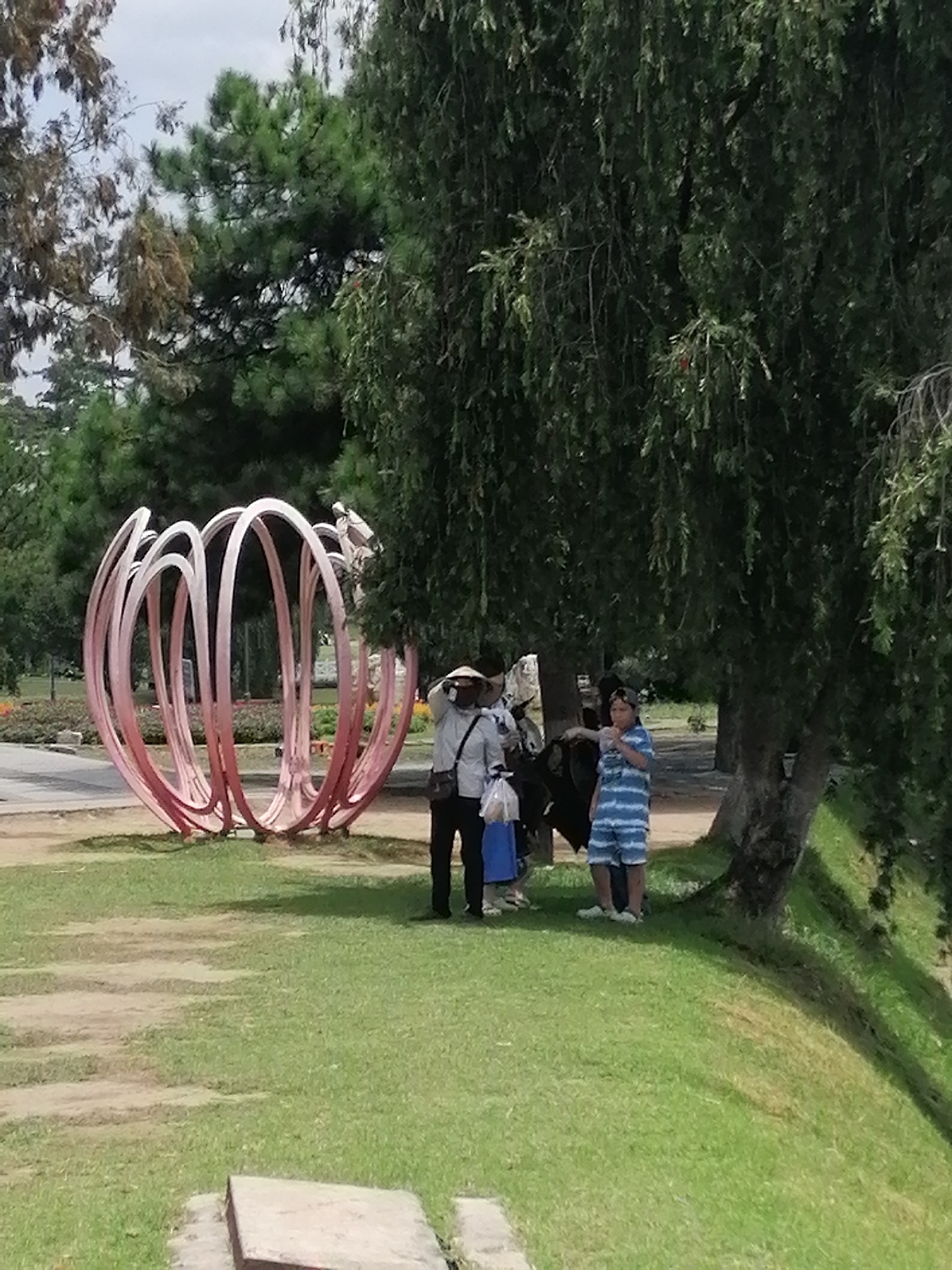 The image depicts a scene in Da Lat, Vietnam, likely at the Da Lat Flower Garden, a popular tourist attraction. In the foreground, a large, abstract sculpture made of pink metal rings stands on a patch of green grass. To the right of the sculpture, under the shade of a large tree, a group of people are gathered. Two individuals, one wearing a conical hat and carrying a bag, and another person in a striped blue and white outfit, are conversing. A small child, also in striped attire, is standing near them. Behind them, a dark shape, possibly a cow or water buffalo, is partially visible. The background reveals more lush greenery, suggesting a park-like setting. The weather appears to be overcast, with a bright sky visible through the leaves of the trees.