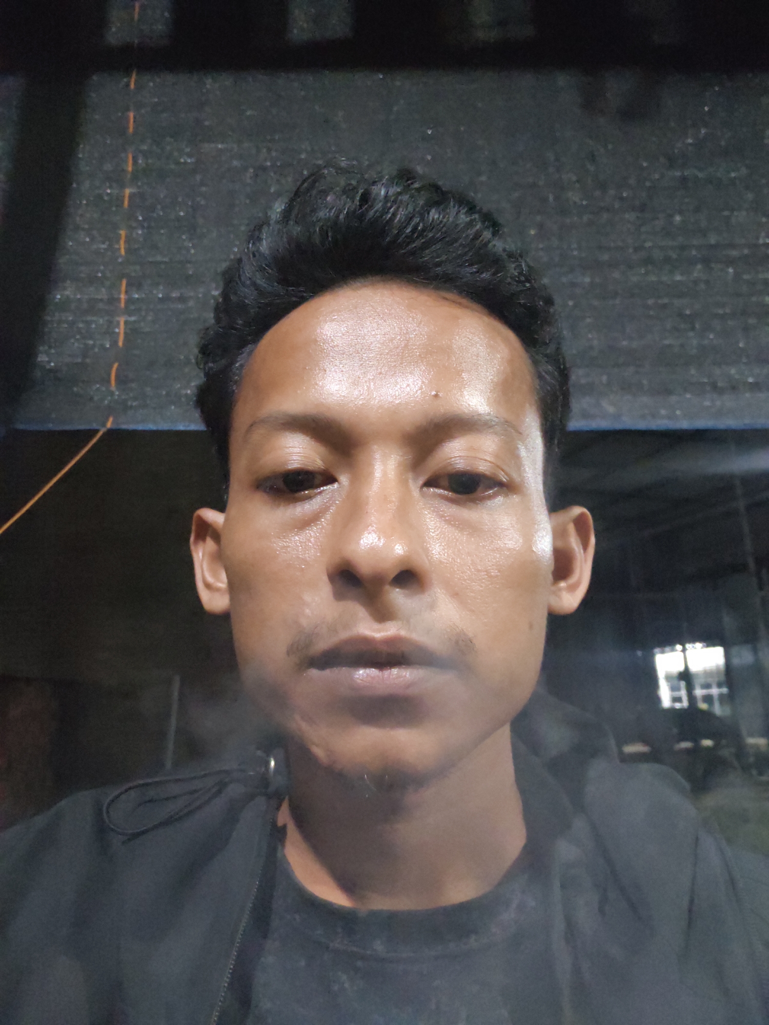 A close-up, head-and-shoulders selfie of a young man is captured in a dimly lit setting. He is wearing a black hooded jacket over a dark t-shirt. His dark hair is styled upwards. His face is illuminated, highlighting his skin which appears slightly sweaty or oily, and his features including his eyes, nose, and lips. He has a faint mustache and stubble on his chin. The background is dark and out of focus, with some structural elements and what appears to be a grate or mesh pattern visible above him. To the right, there are some illuminated rectangular shapes, possibly windows or lights, suggesting an indoor or covered outdoor space. The lighting suggests it could be nighttime or a dark indoor environment. No visible text is present in the image. The overall impression is a casual, informal self-portrait.