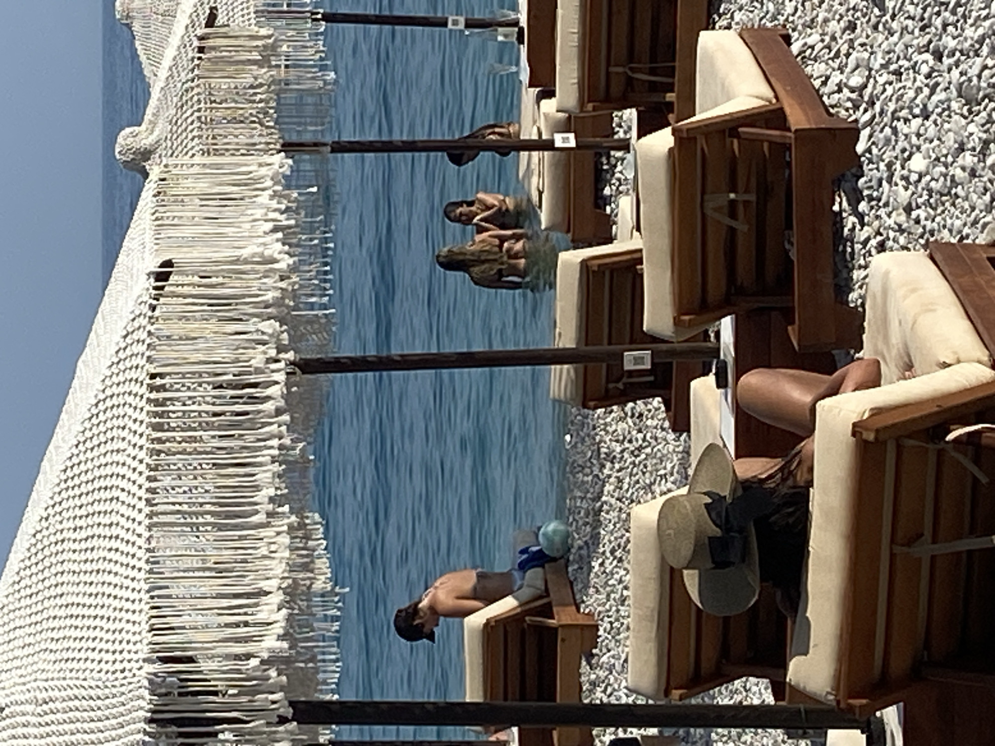 This is a medium-angle, eye-level, full shot of a beach scene in Prijevor, Montenegro. The foreground is dominated by several wooden lounge chairs with beige cushions arranged on a pebbled beach. A person wearing a wide-brimmed hat is seated in one of the chairs, facing away from the camera, with their legs extended. The chairs are positioned under large, white, macrame-style umbrellas with fringe. In the middle ground, the clear blue water of the Adriatic Sea is visible. Several people are in the water; one person is sitting on a lounge chair at the water's edge, facing the sea. Further out in the water, three people are wading, two facing each other and talking, and a third person is further to the right. The background shows a clear blue sky and the horizon where the sea meets the sky.