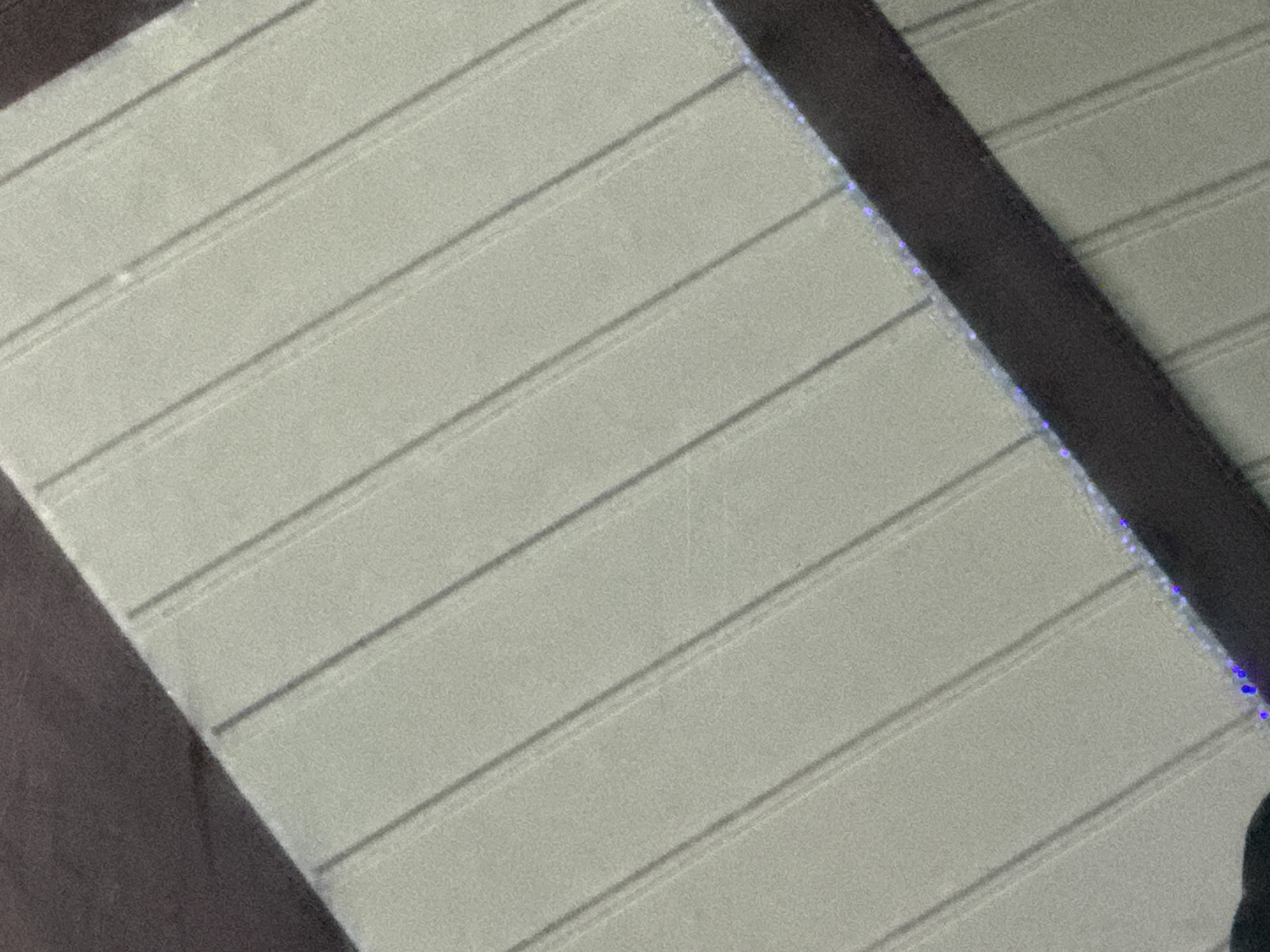 The image displays a close-up, angled view of a light gray, textured surface with parallel vertical indentations or ridges. The surface appears to be made of a flexible material, possibly fabric or a similar synthetic. The indentations are evenly spaced and run diagonally across the frame. A dark, solid band, appearing to be a strap or border, runs diagonally across the lower portion of the image. This band is adorned with small, bright blue lights or reflections along its edge. A dark, blurry area is visible in the upper left corner, possibly indicating an object or shadow outside the primary focus. The overall scene is indistinct in terms of specific location, and no elements within the image suggest a connection to Slahung, Indonesia, or any other specific urban environment.