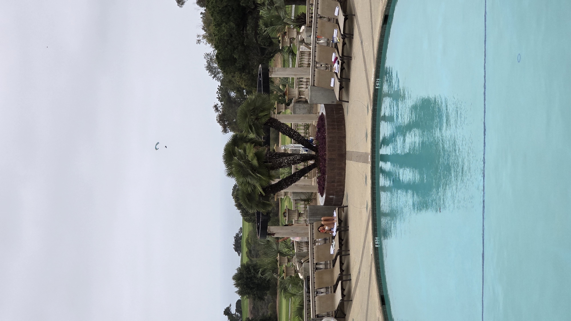A person is reclining on a lounge chair by a swimming pool, appearing to eat a piece of fruit. The pool area is paved and features several other empty lounge chairs, each with towels. A circular planter with palm trees stands prominently in the background, with an ornate stone balustrade and columns surrounding it. Beyond the pool area, a golf course is visible, with rolling green hills and trees under a cloudy sky. In the distance, a paraglider is visible in the sky. The image appears to be taken in La Jolla, San Diego, United States of America.