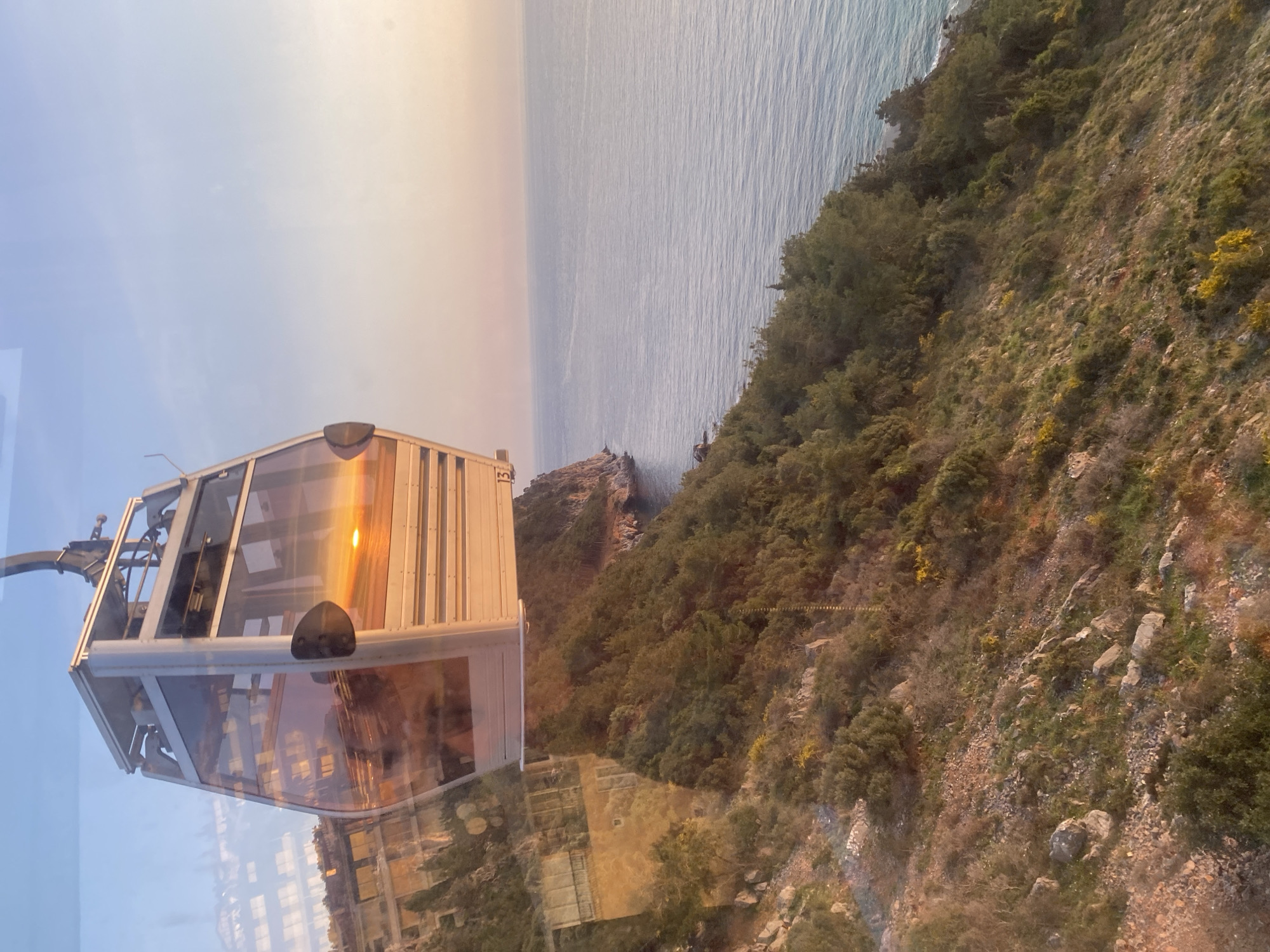 The image shows a cable car in Alanya, Turkey, with a view of the coastline. The cable car is predominantly white and has large glass windows. The reflection of the sunset or sunrise is visible on the windows. The terrain outside the cable car slopes downwards and consists of greenery and rocks. The Mediterranean Sea is visible in the background, meeting the horizon under a blue sky. The light from the sun gives a warm glow to the scene.