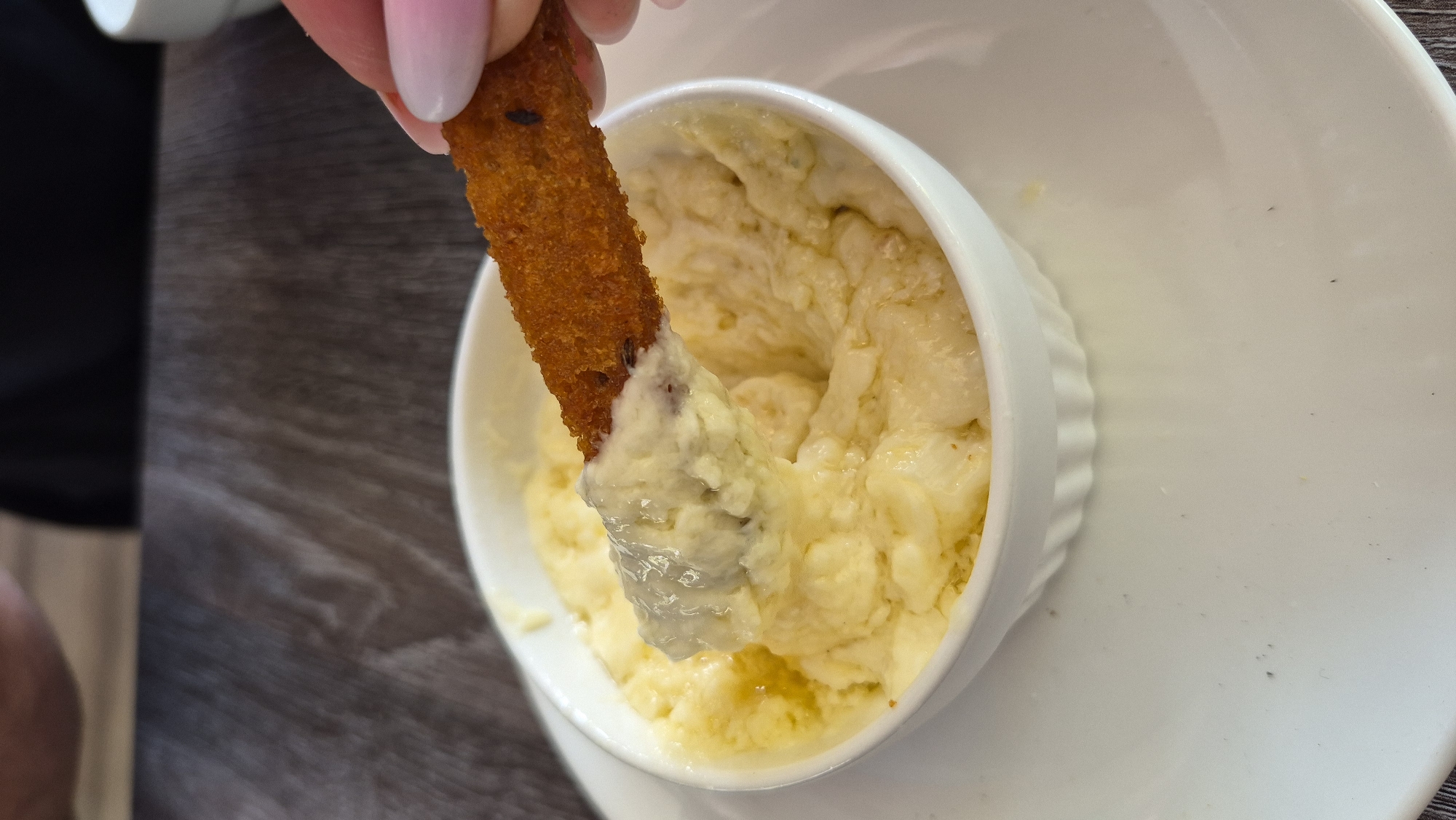A close-up shot shows a person's hand dipping a piece of fried food into a bowl of creamy, yellow dip. The dip appears to be a cheese-based spread, possibly fondue or a similar appetizer. The fried piece is golden brown and textured, suggesting it's a breadstick or a similar crunchy component. The bowl is white and rests on a white plate. In the background, a dark wooden table is visible, and the blurred figures of people suggest a restaurant or social setting. The overall lighting is bright, highlighting the food. This scene is likely taking place in a restaurant in Zarasai, Lithuania, given the context provided.