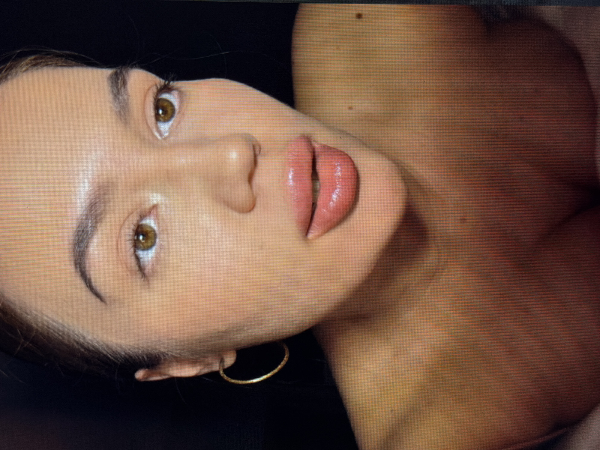 The image is a close-up, low-angle shot of a woman's face and upper chest, with a dark background. The woman has brown eyes, full lips with pink lipstick, and is wearing a gold hoop earring. Her skin appears to have a light foundation or makeup applied. Her hair is pulled back, and part of her décolletage is visible. There are a couple of small dark moles on her upper chest. The image quality suggests it was taken on a digital screen, possibly a monitor or TV. The description of the location in Sirius, Russia, is not supported by any visual evidence in the image itself.