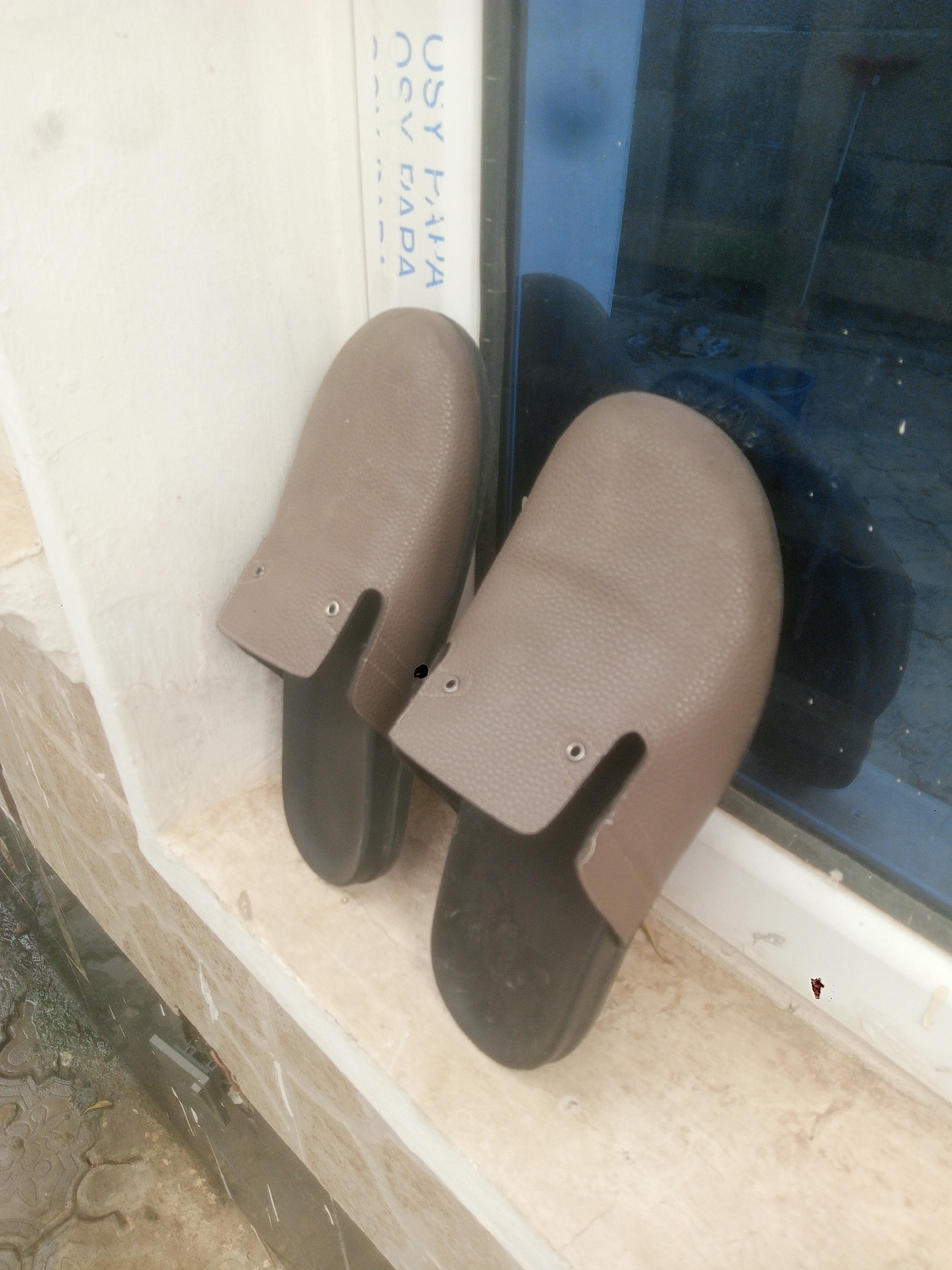 Two brown leather-look sandals rest on a beige windowsill next to a window. The sandals are open-toed slip-ons with visible black soles and small metal eyelets near the top of the upper portion. The windowsill is bordered by a white wall. Outside the window, reflections and outdoor objects are visible. The location is Kubwa, Nigeria, indicated by the local brand name "USY PAPA OSV DADA" printed on the window frame.
