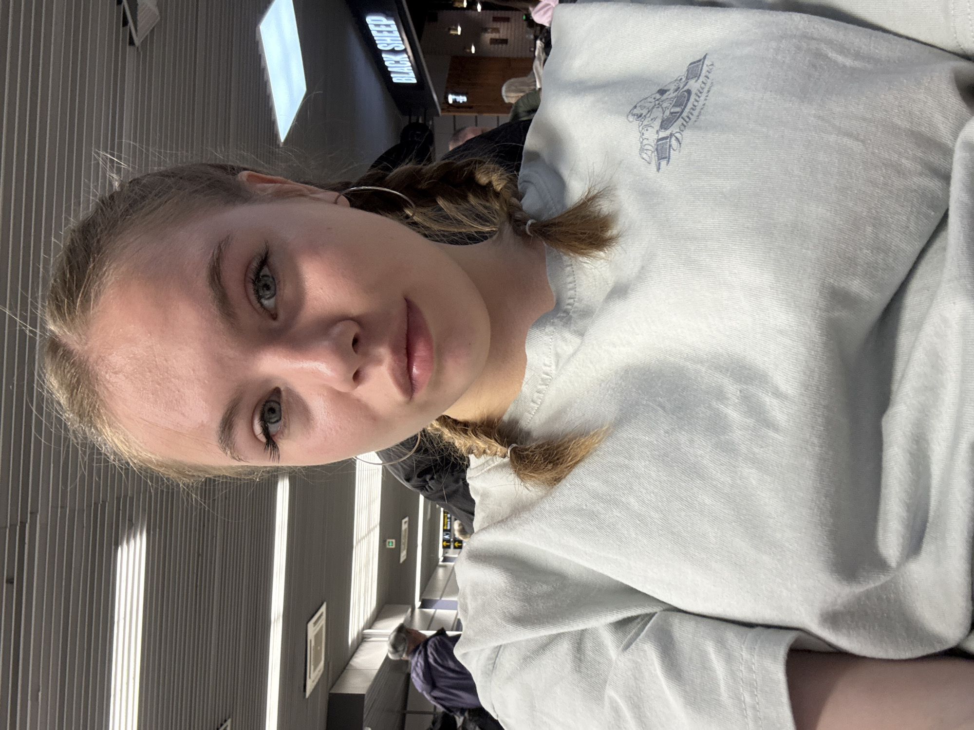 A young woman with blonde hair braided into two pigtails is pictured in an indoor setting, likely an airport terminal based on the background elements. She is wearing a light-colored t-shirt with a graphic on the shoulder. The background reveals a hallway with a 'Black Sheep' sign, various figures walking around, and architectural details such as wall panels and signage. The location is indeterminate and cannot be confirmed as Sochi, Russia, without additional context or landmarks.