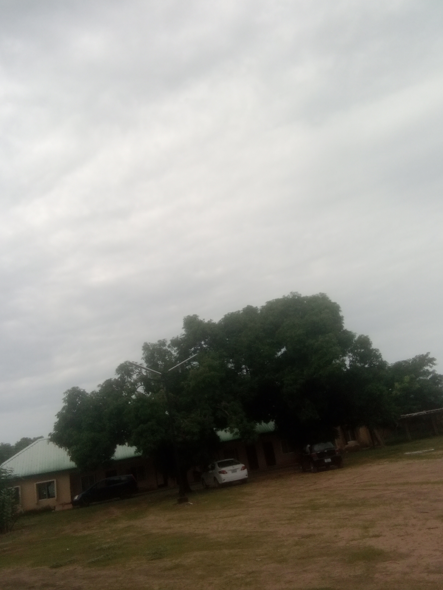 The image shows a building with a green roof and several cars parked in front of it. The building appears to be a single-story structure with windows and doors. There are trees and grass around the building, and the sky is overcast. The scene is likely located in Jalingo, Nigeria, given the vegetation and architectural style.
